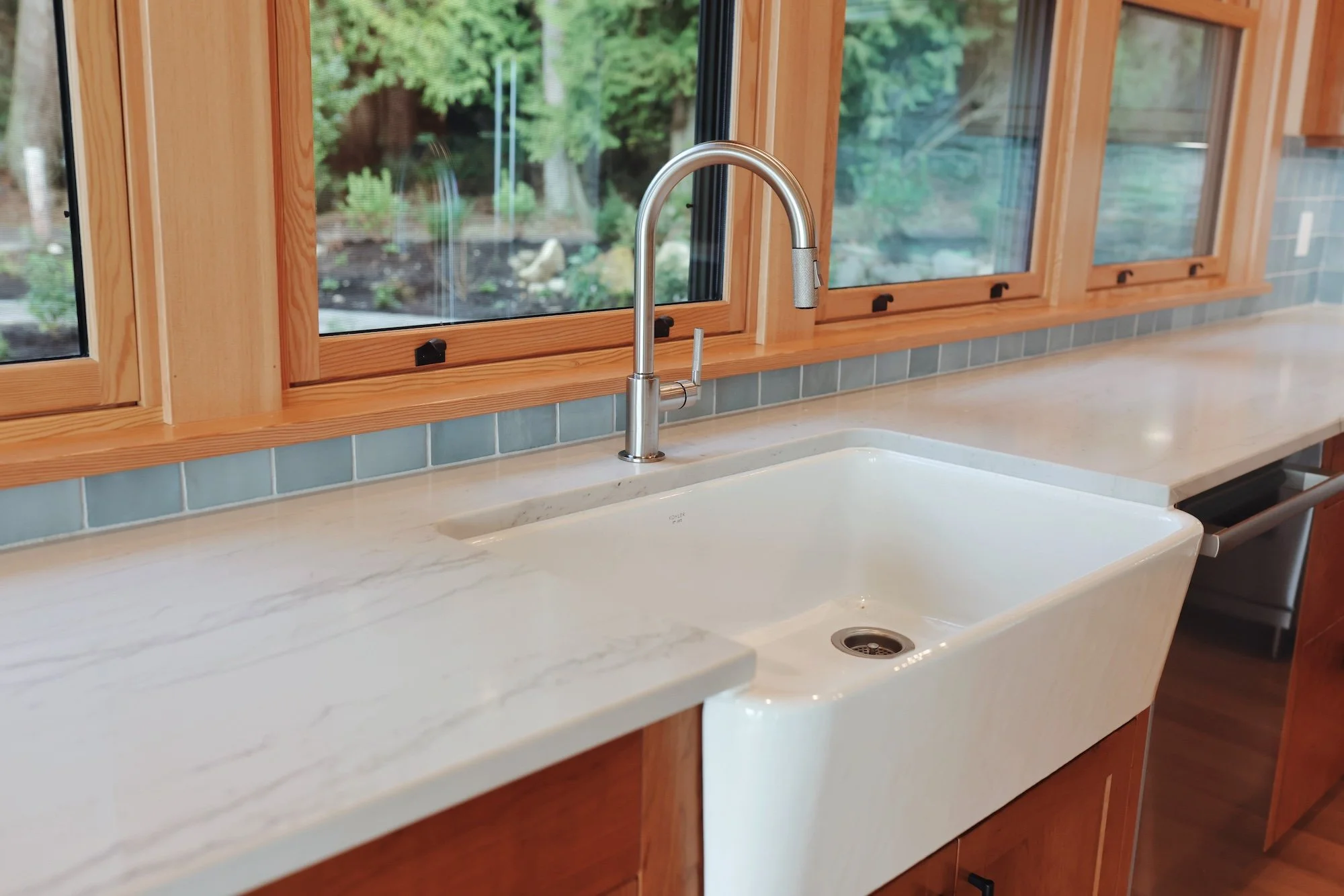 Indoor kitchen with white marble countertop, stainless steel faucet, and large wooden framed windows overlooking a garden.