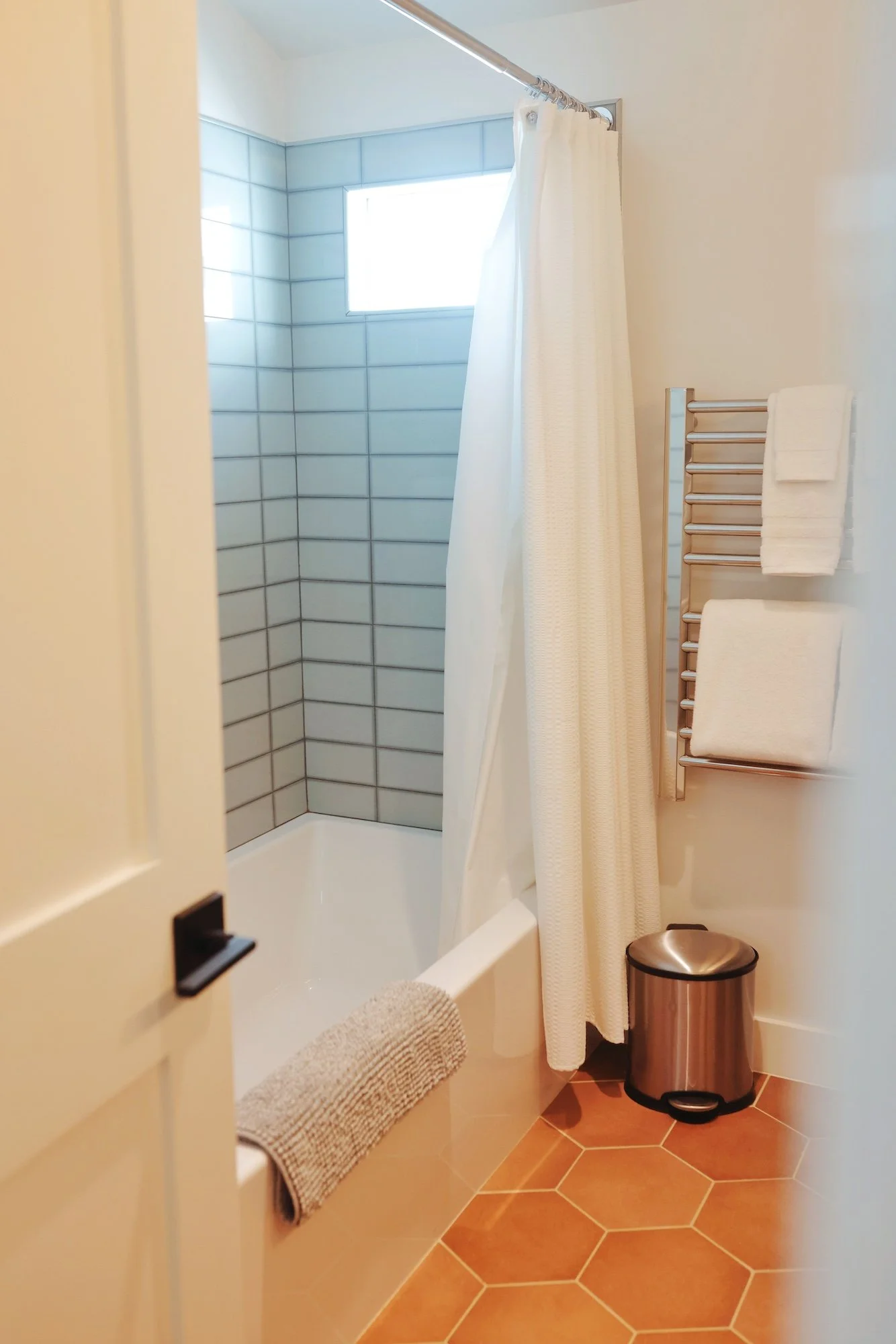 A bathroom shower with white walls, light blue tiles, a window, a white shower curtain, a towel rack with towels, a small trash can, and a beige bath mat on the terracotta tiled floor.