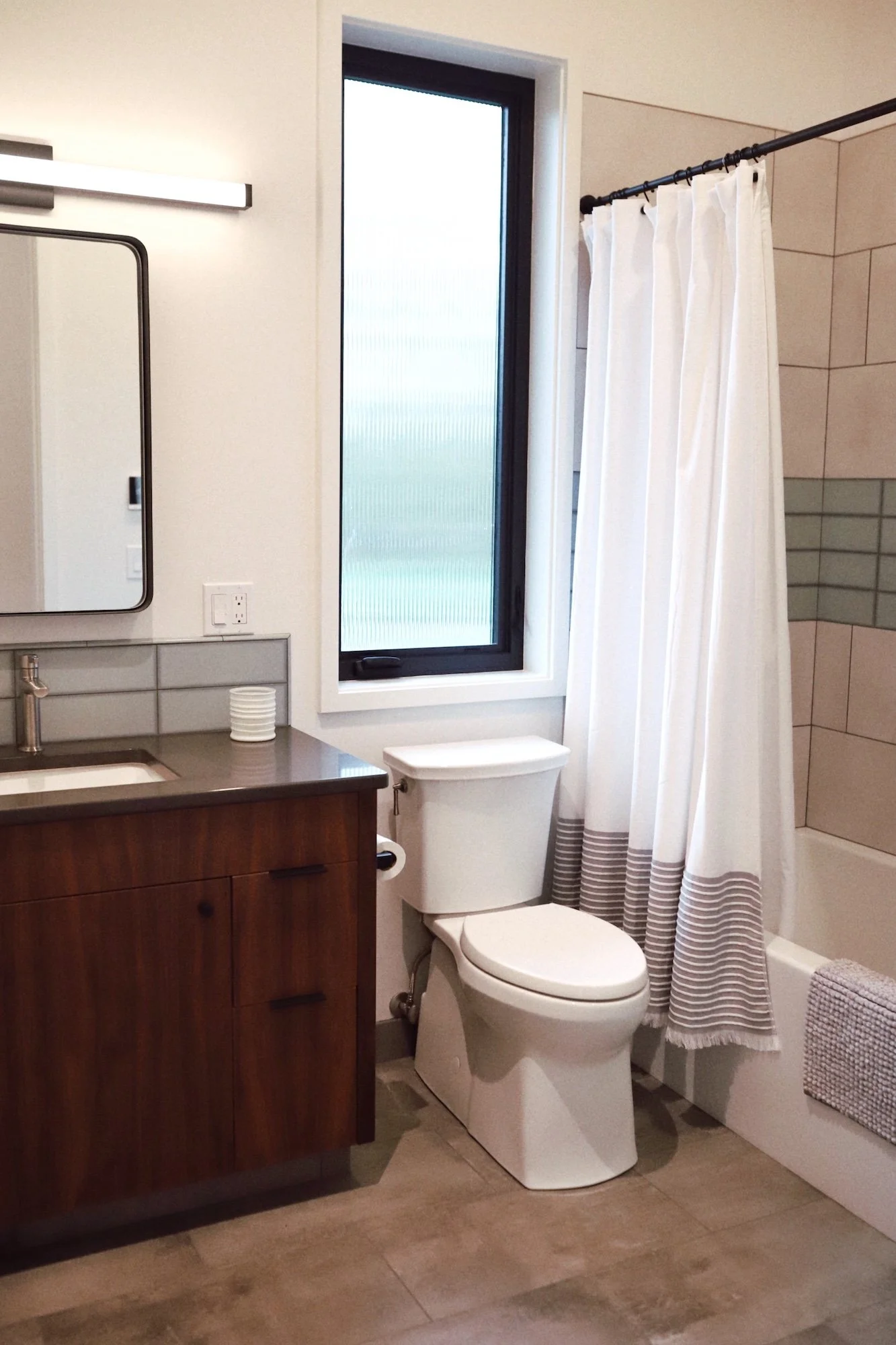 Modern bathroom with a wooden vanity, mirror, white toilet, window, and shower with a striped curtain.