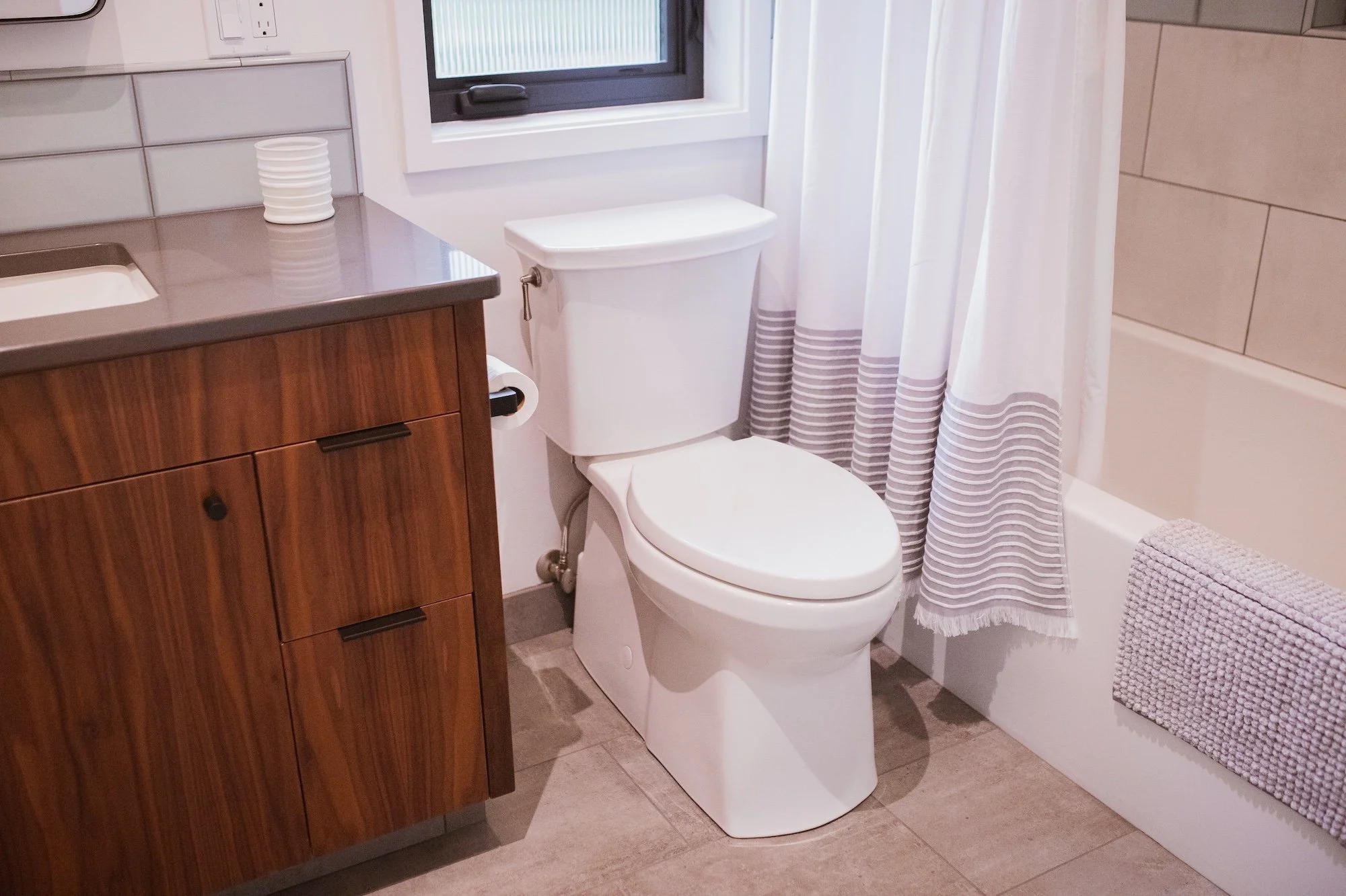 A bathroom with a toilet next to a wooden cabinet, a window with blinds, and a shower curtain with striped pattern.