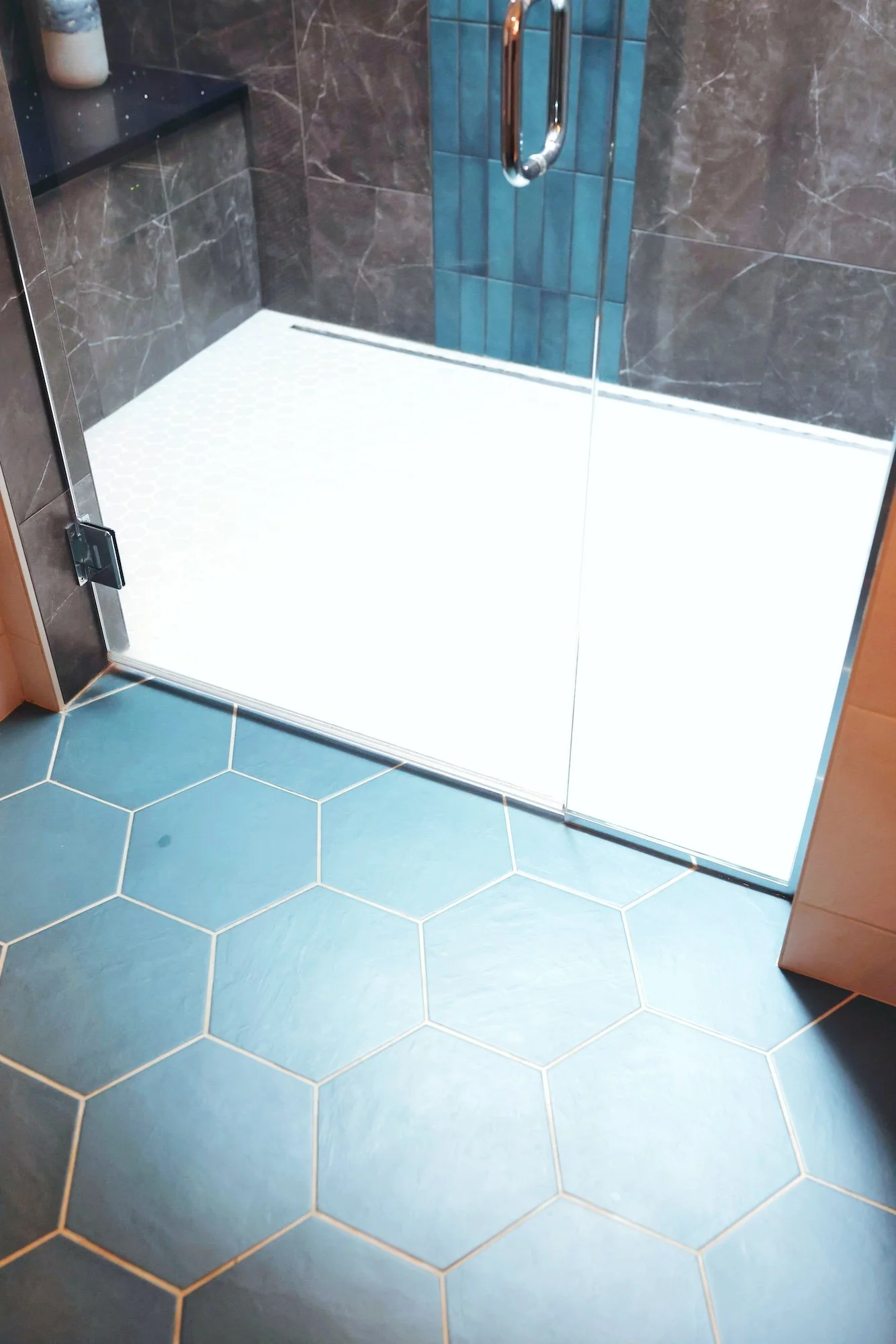 View of a shower with tile walls and blue hexagonal floor tiles, glass door, and a metallic handle.
