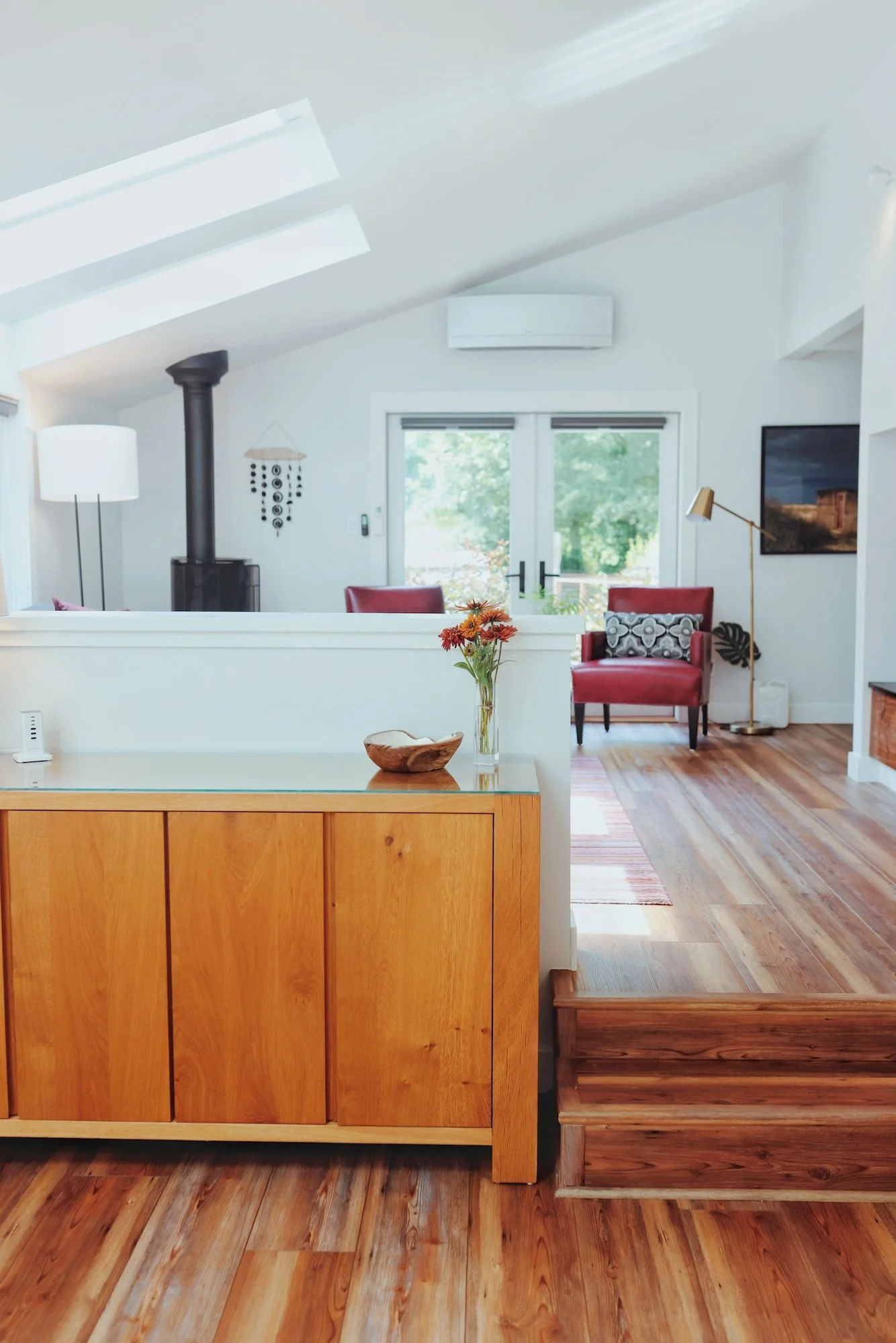 Living room with hardwood floors, white walls, and large sliding glass doors leading outside. Contains a black wood stove, a tall white floor lamp, a red armchair with a black and white cushion, a brass floor lamp, and artwork on the wall. A small wo