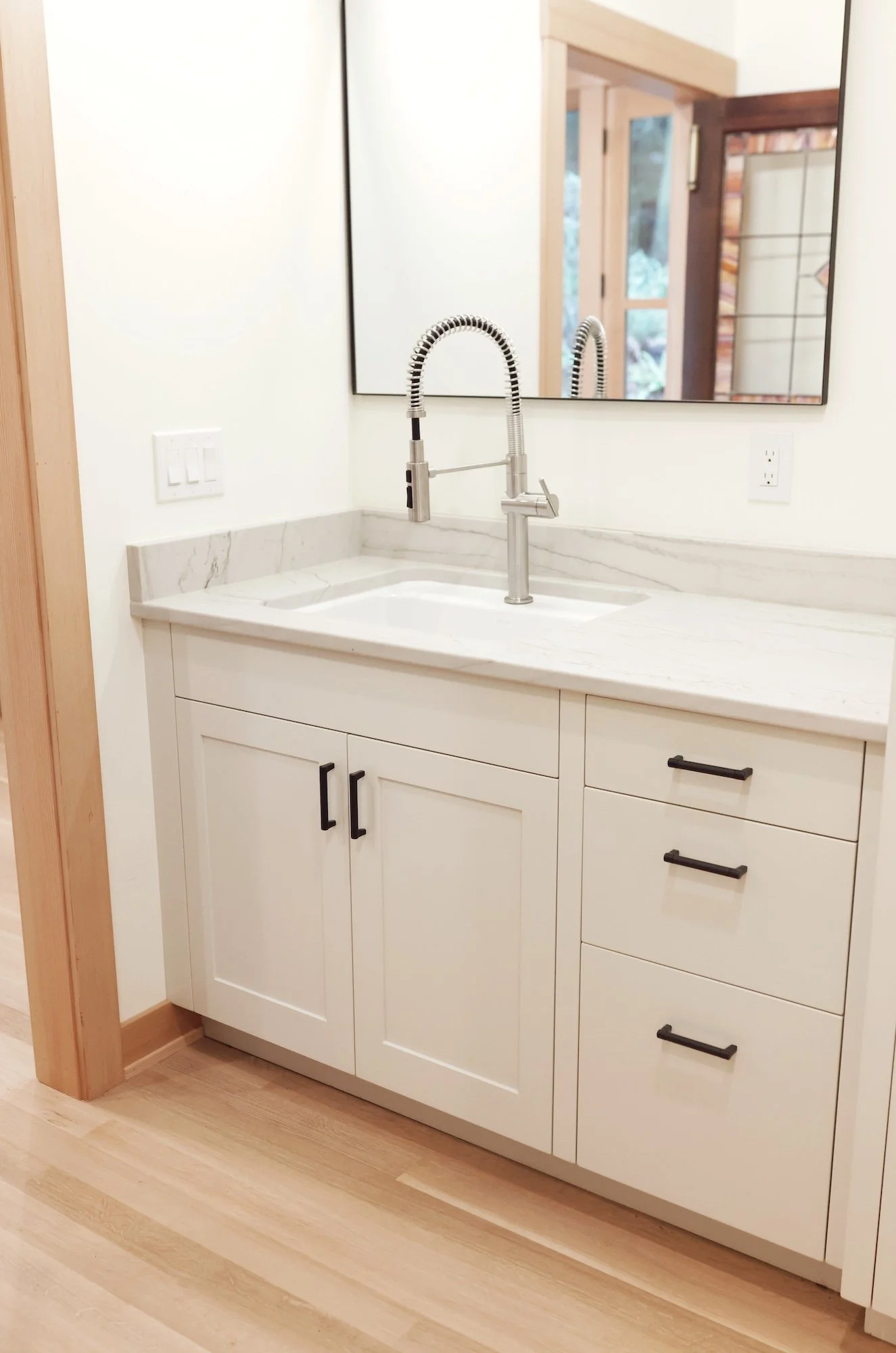 Close-up of a modern kitchen sink with a chrome faucet, white cabinetry, and a marble countertop. A large mirror and a window reflecting interior details are in the background.