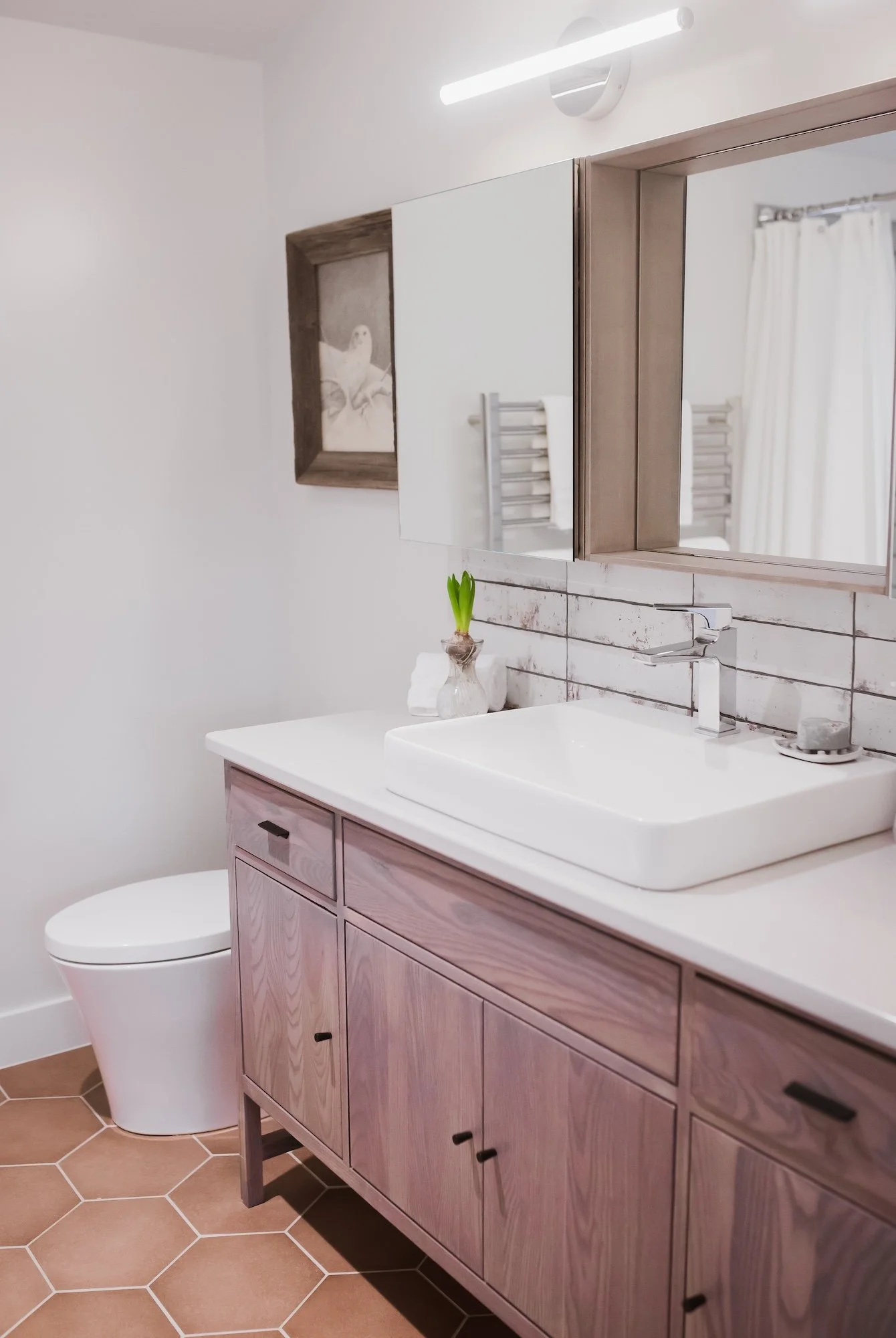A modern bathroom with a white sink, wooden vanity, white toilet, and a large mirror. There is a small potted plant on the sink. The mirror is framed with wood and a framed artwork of an owl is on the wall. The bathroom has white walls, a light fixtu