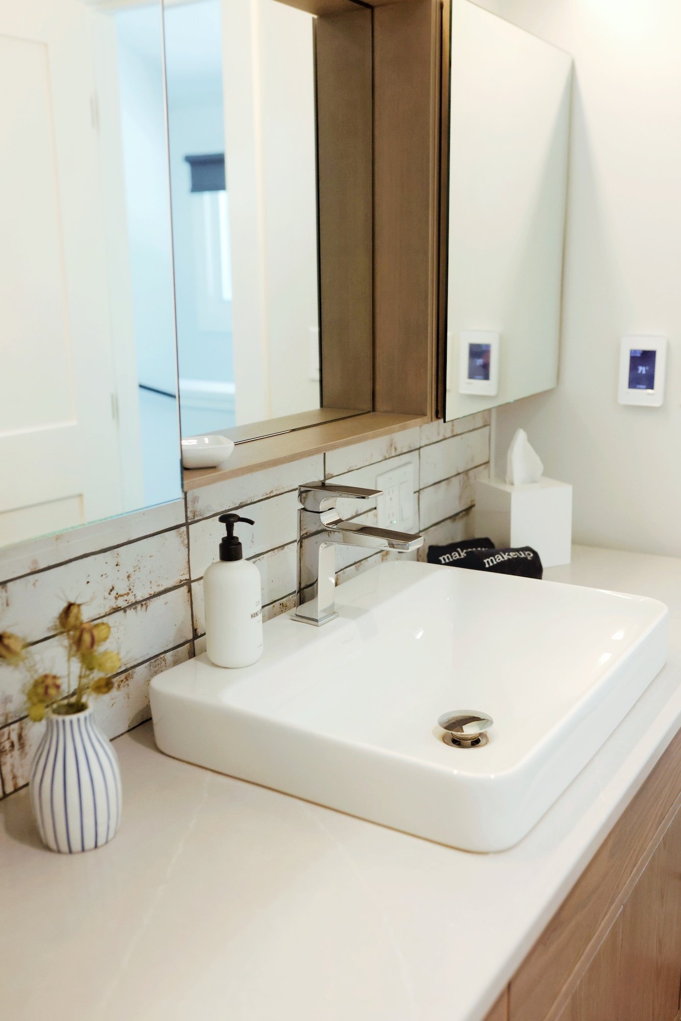 Modern bathroom with white rectangular vessel sink, gold faucet, and white wall-mounted thermostat. A small vase with dried flowers and a soap dispenser are on the sink's counter. Mirror above the sink reflects part of the bathroom. The wall behind t