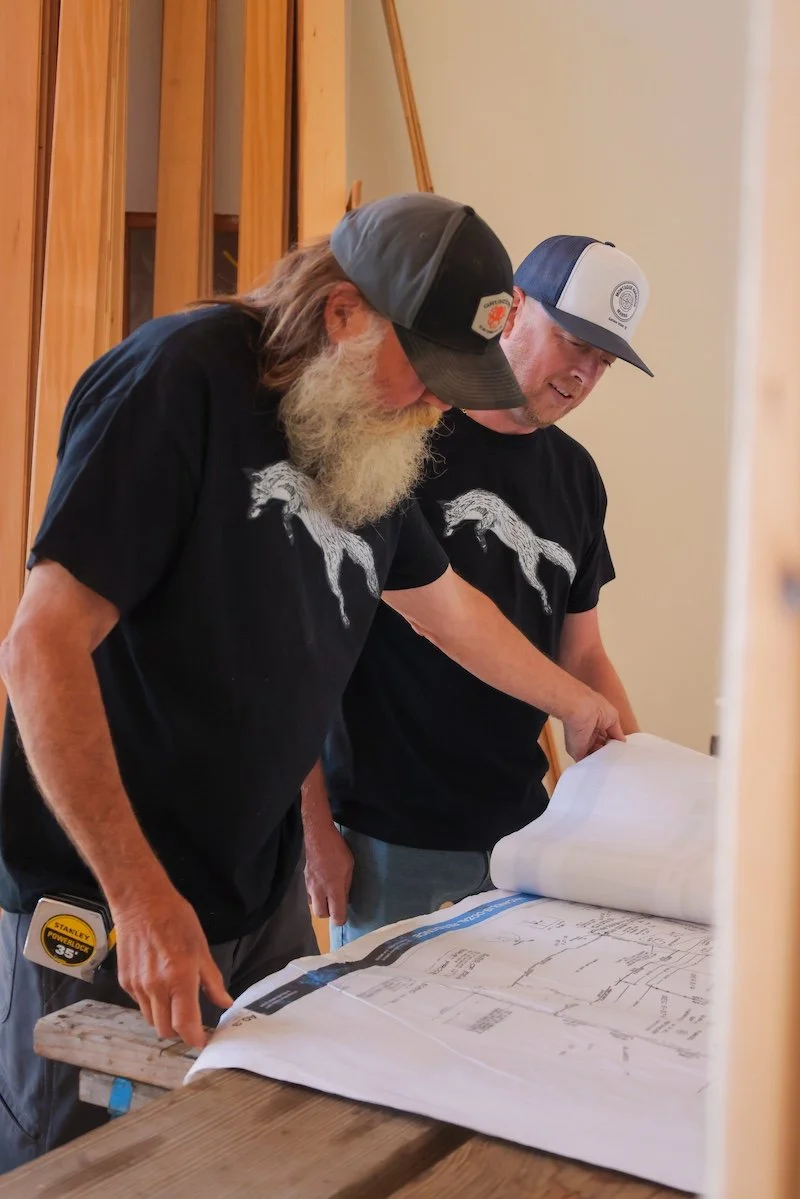 Two men wearing black t-shirts with a horse graphic are looking at construction plans on a table, inside a building under construction with wooden framing.