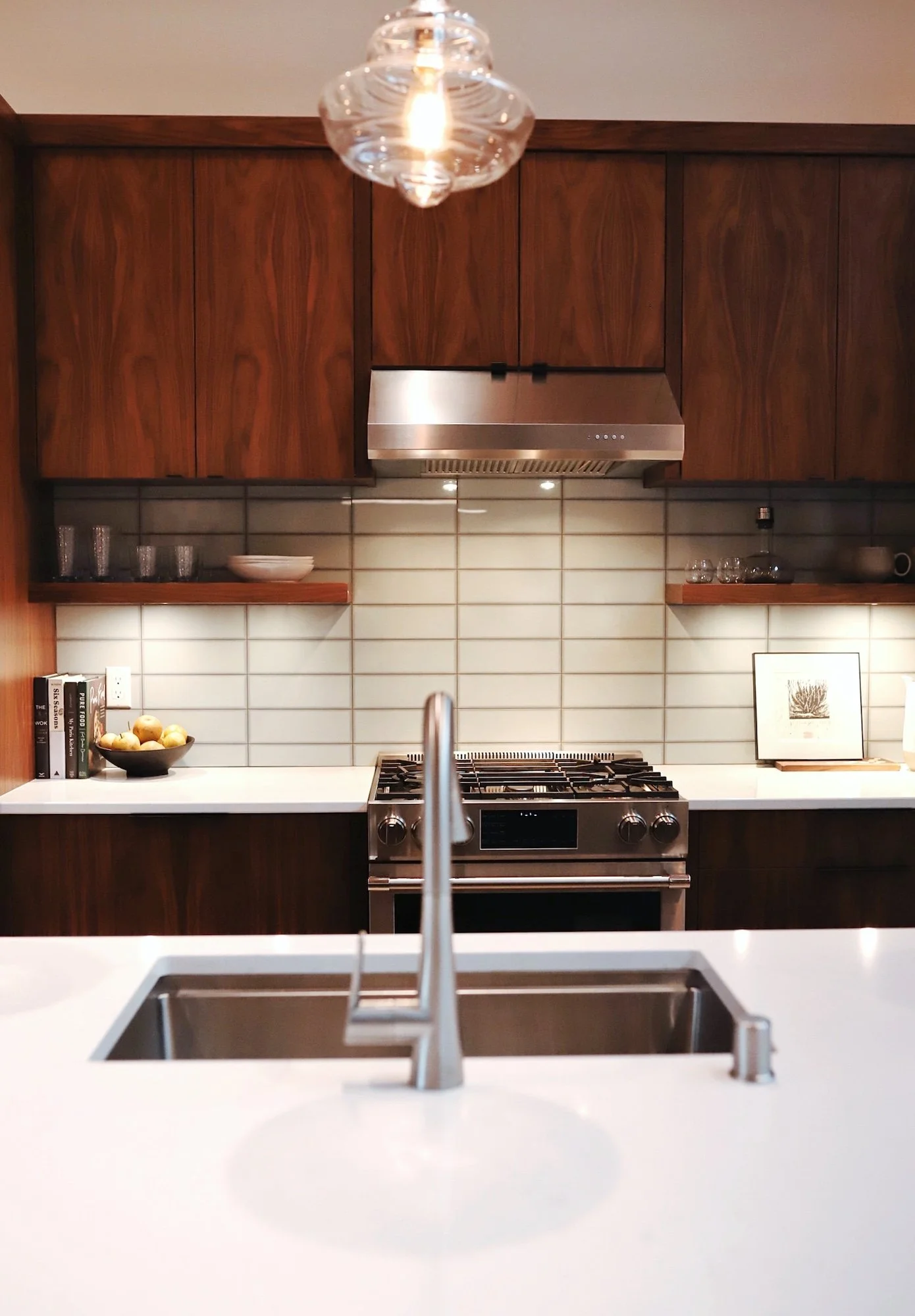 Modern kitchen with dark wooden cabinets, a stainless steel range hood, white tile backsplash, and a white countertop with a black bowl of apples. The photo is taken from the perspective of the kitchen sink with a stainless steel faucet visible in th