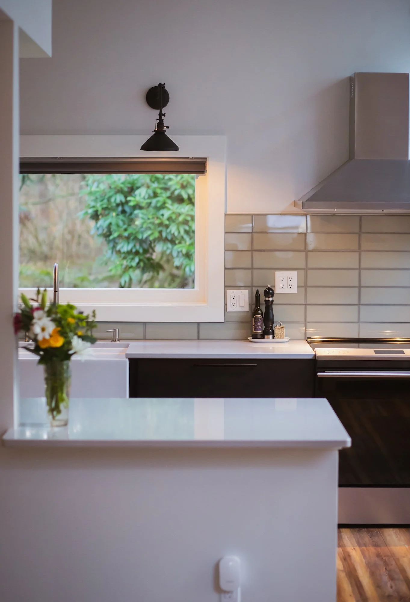 Modern kitchen with a window showing green foliage outside, a white countertop with a vase of flowers, and black utensils next to a stovetop. The wall has beige tiles, and there is a stainless steel range hood and electrical outlets.