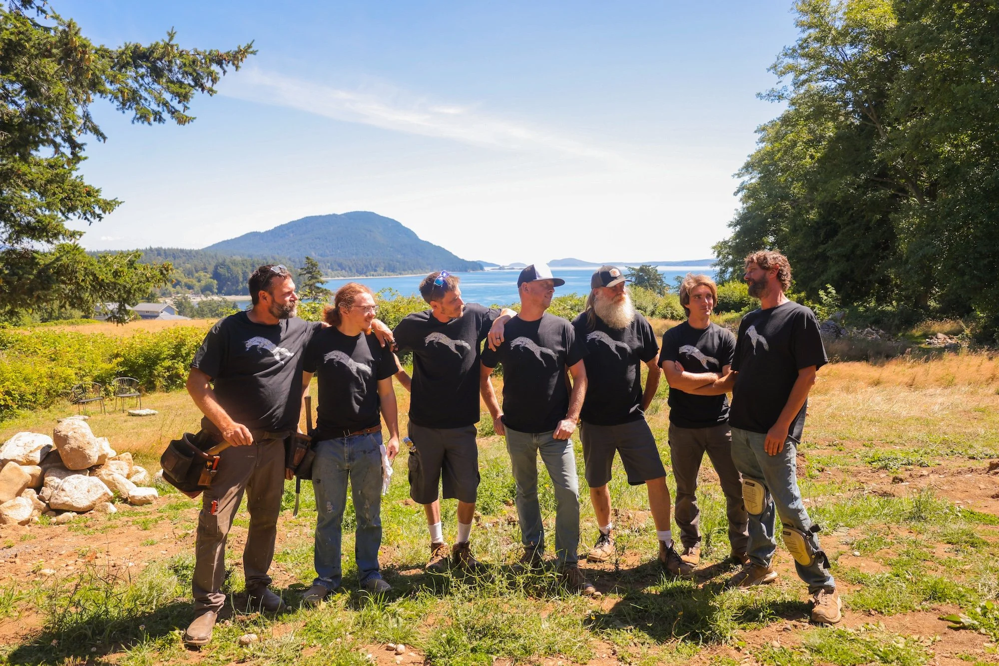 Group of seven people standing outdoors on a farm or park, with a lake and mountains in the background, under a clear blue sky, engaged in conversation and smiling.