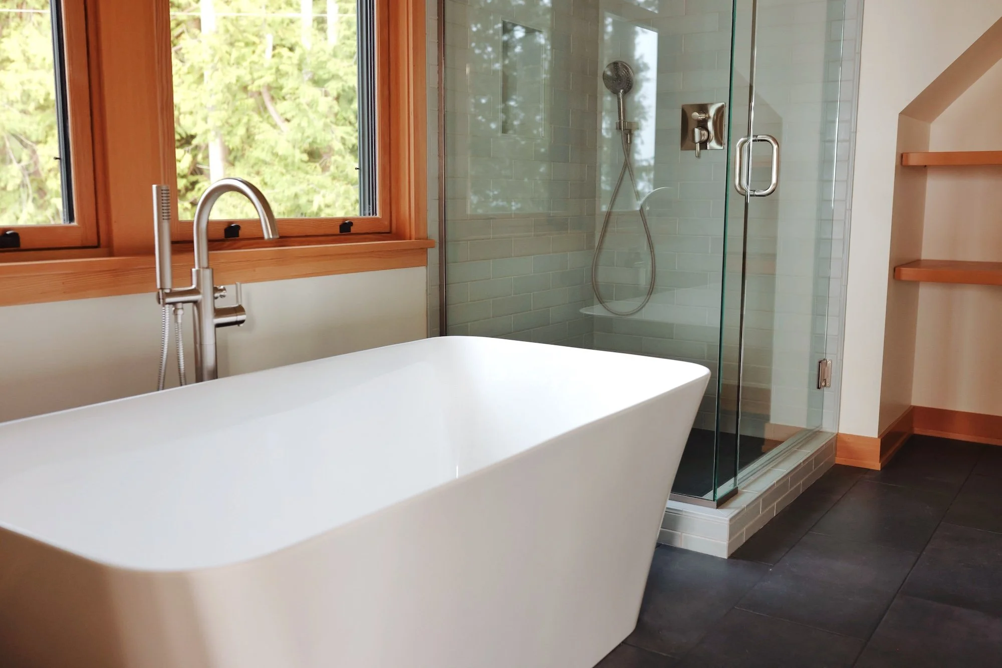 Modern bathroom with a freestanding bathtub next to a large window, a glass-enclosed shower with a showerhead and controls, and wooden shelves on the wall.