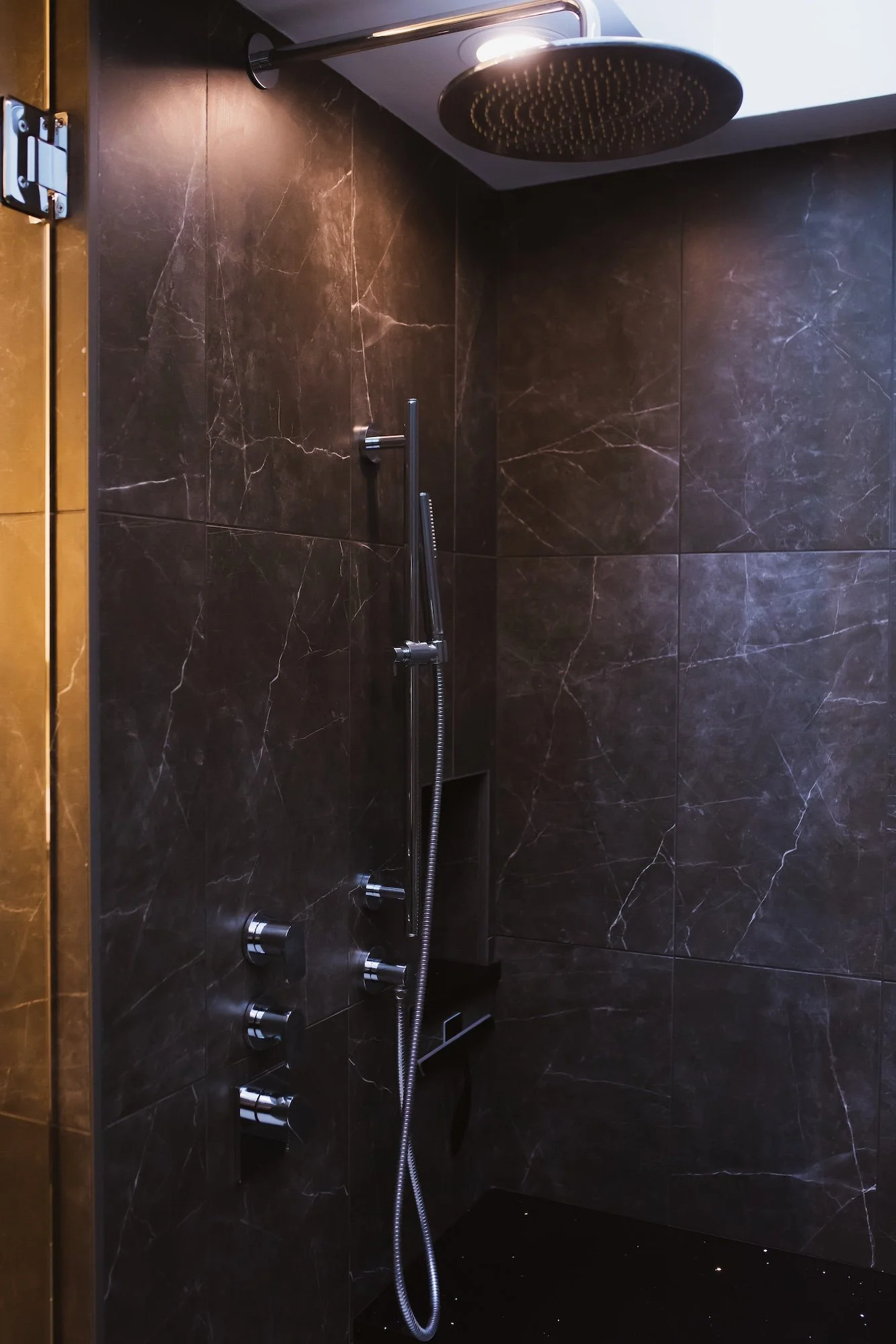 Inside a modern shower with dark marble tiles, a rain showerhead, a handheld shower wand, and control knobs.
