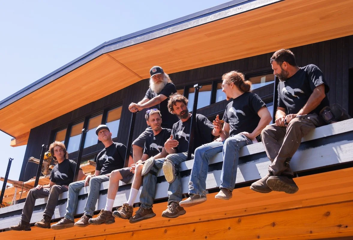 Seven people sitting on a wooden balcony, some talking and one with a beard and sunglasses. They are outside a modern black house with large windows and a sloped roof, sunny day.