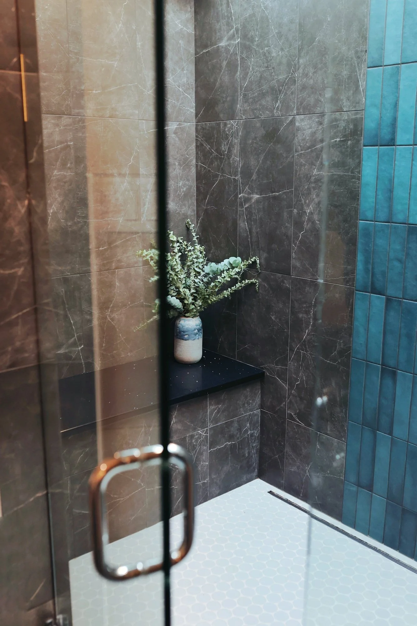 A view of a modern bathroom shower area with dark gray marble tiles on the walls, a small black platform with a potted plant, blue tiles on one side, and white hexagonal tiles on the floor.