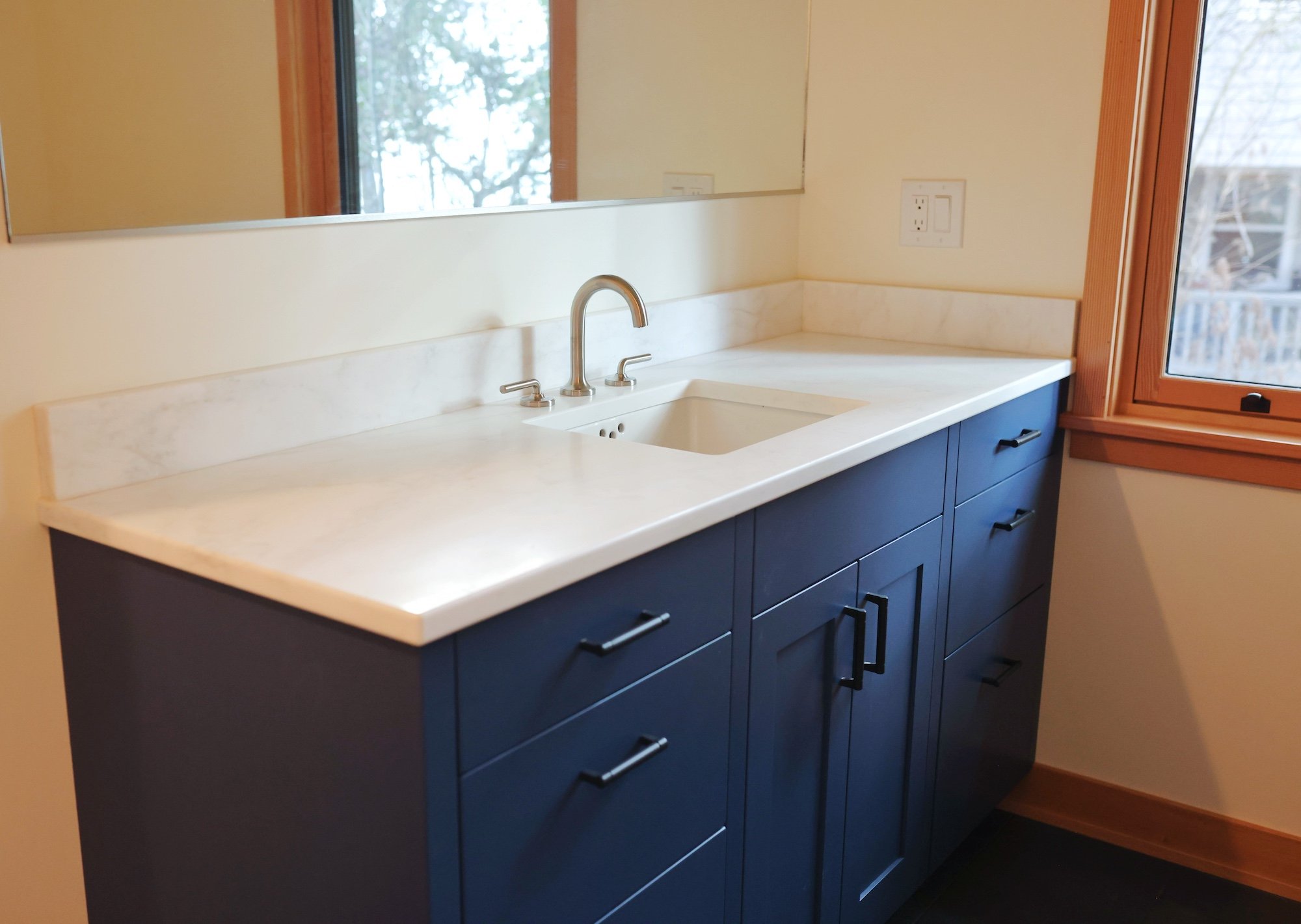 A modern bathroom vanity with a white marble countertop, a rectangular undermount sink, a brushed nickel faucet, and dark blue cabinet drawers with black handles. There are two electric outlets on the wall above the countertop, and a large mirror ref