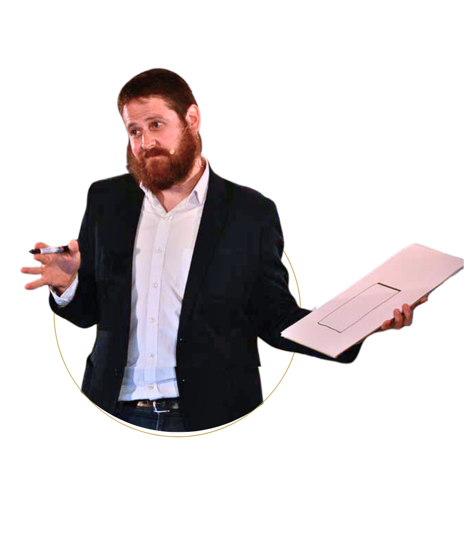 Man with a beard in a suit holding a closed laptop and a marker, speaking in front of a black background.
