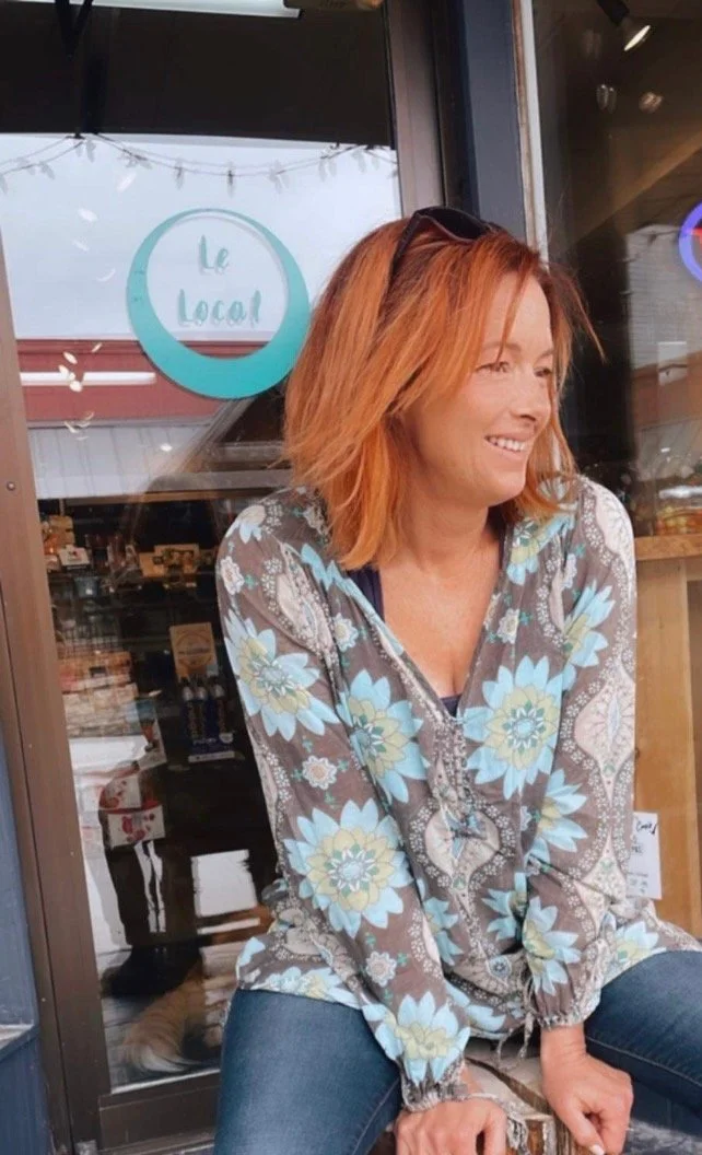 Femme rousse assise devant un café, portant une veste à motif floral, souriant, avec une enseigne 'Le Local' en arrière-plan.