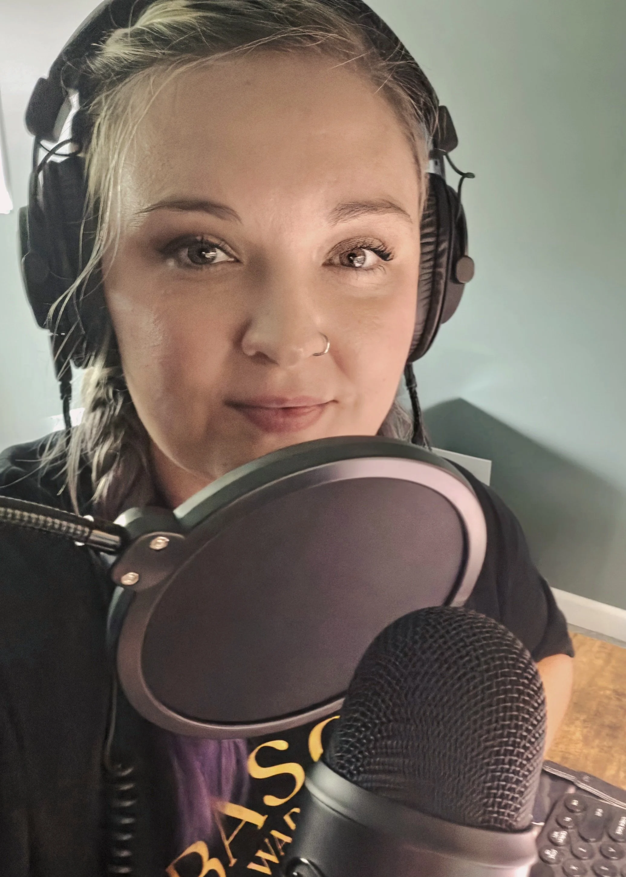 Close-up of a young woman with blonde hair wearing headphones and a nose ring, recording in a studio with a microphone and audio equipment.