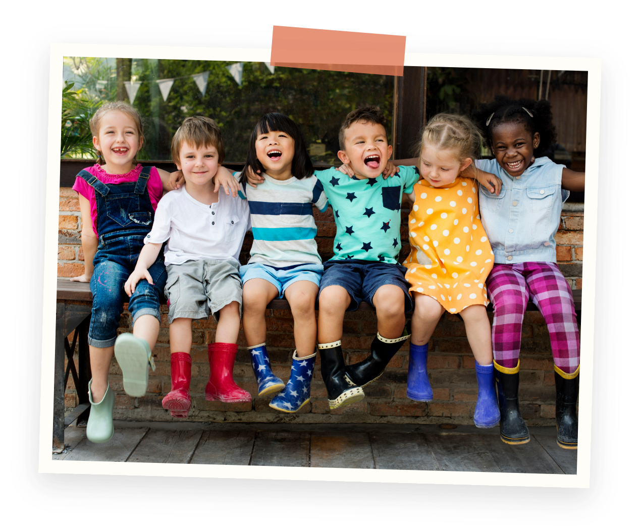 Preschool program kids having fun. Group of seven diverse children sitting on a wooden bench outdoors, smiling and laughing, with hands around each other's shoulders.