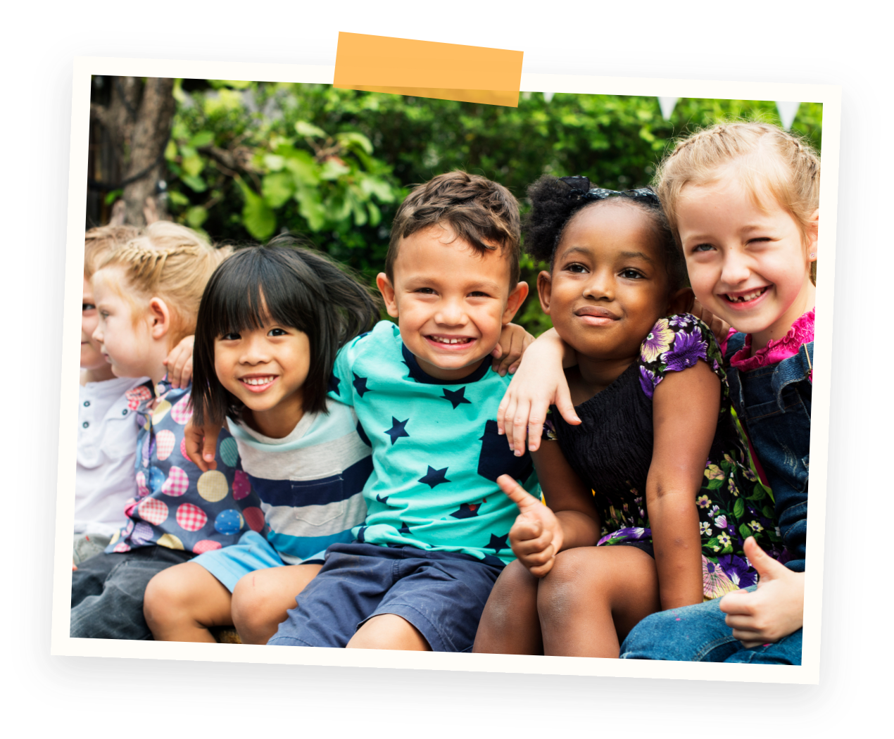 Outdoor play area at our Marpole Preschool. Group of diverse children sitting outdoors, smiling and posing for a photo, with trees and greenery in the background.