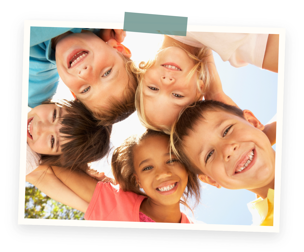 A group of six children smiling and looking down at the camera with their heads together in a circle, outdoors on a sunny day.