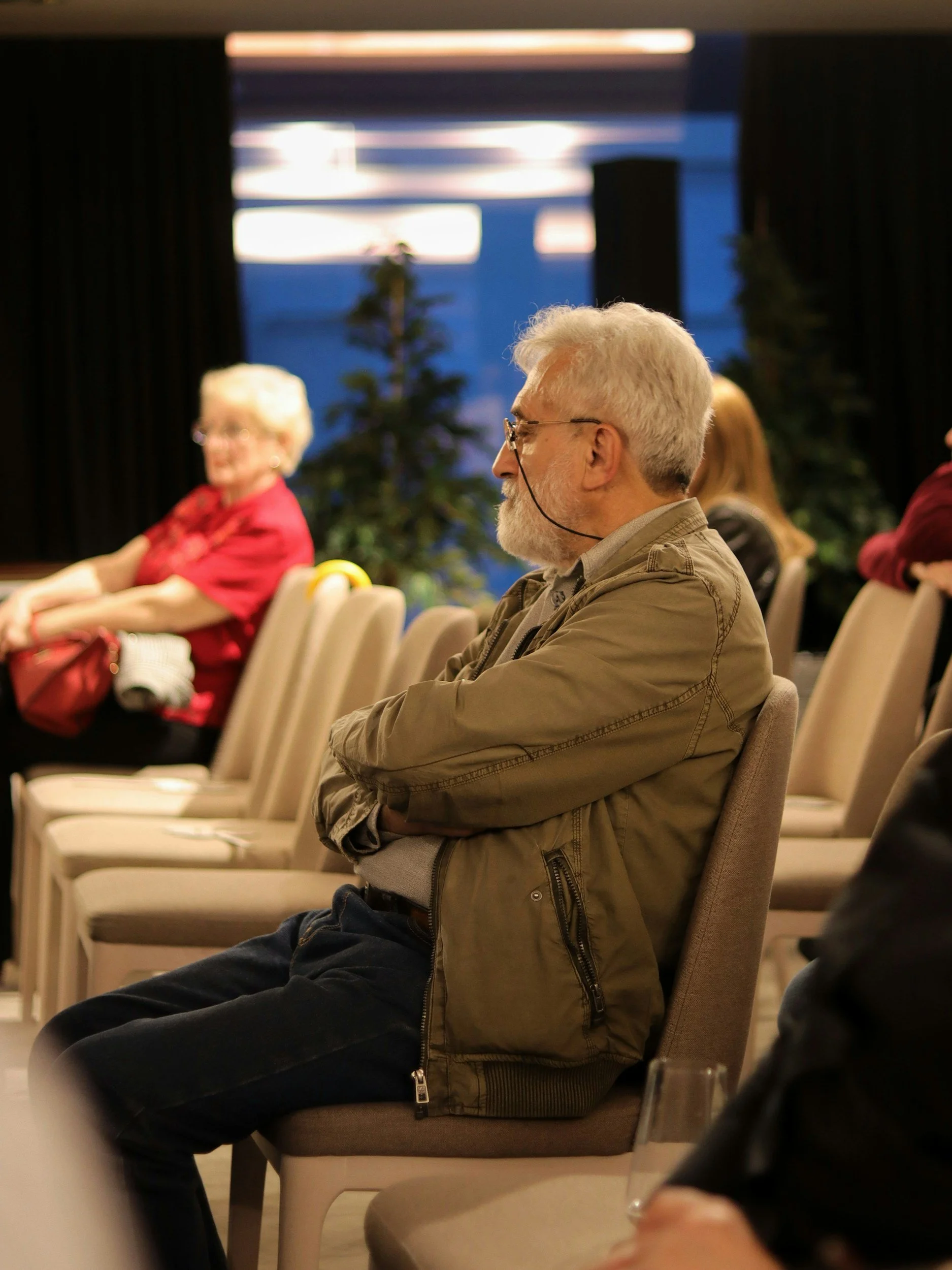 A group of people seated in a room, attending an an owners' meeting, with focus on an older man with gray hair and beard, wearing glasses and a brown jacket.