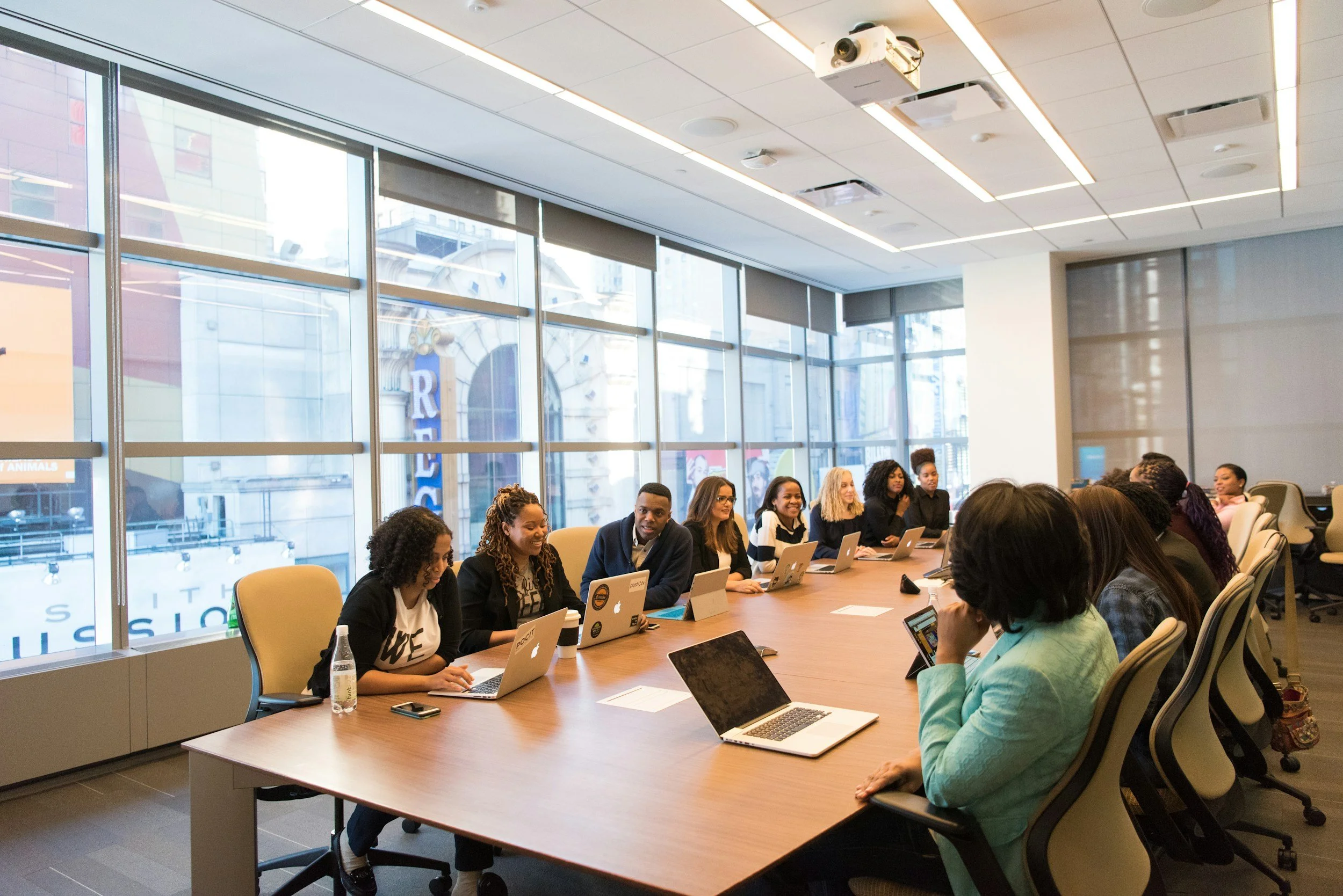 A diverse group of people sitting around a large conference table in a modern office, working on laptops, with large windows and city buildings visible outside.