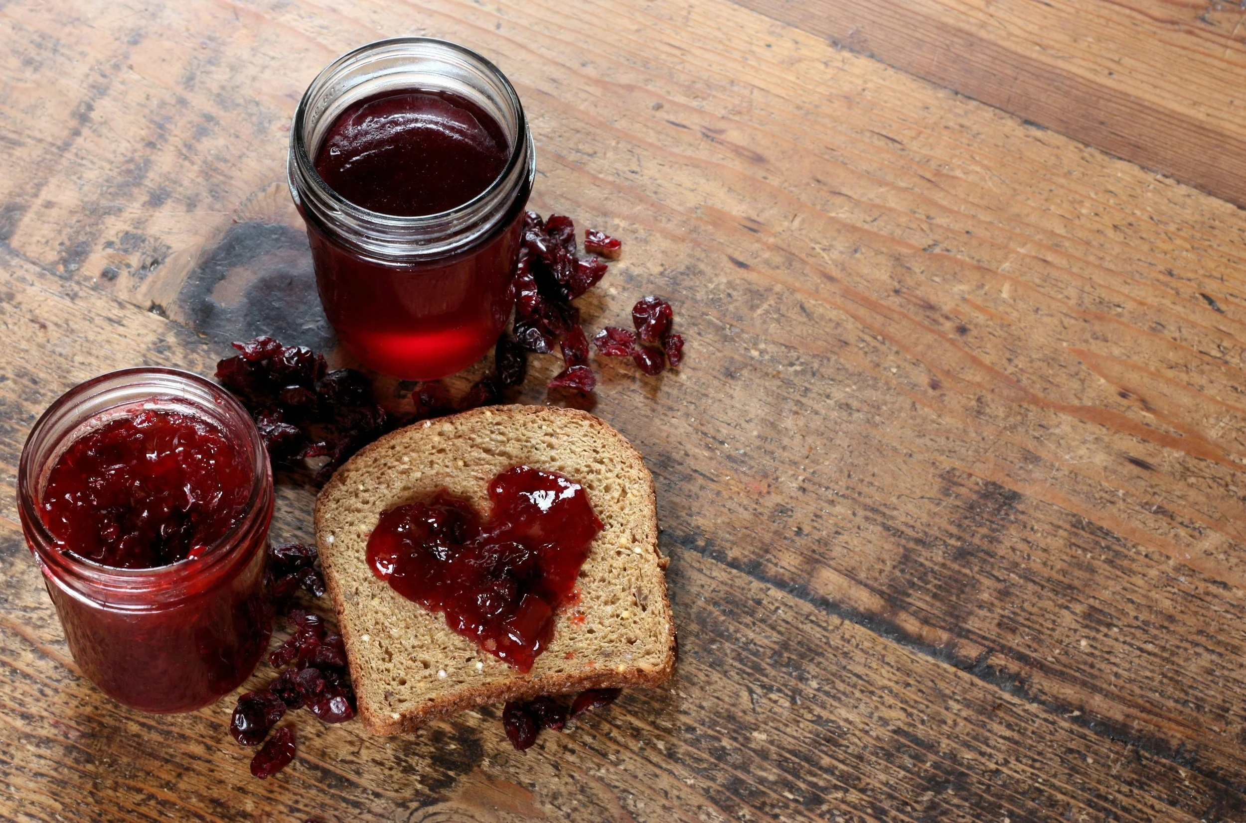 A jar and a glass of dark red berry jam on a rustic wooden table, with a slice of bread spread with jam nearby.