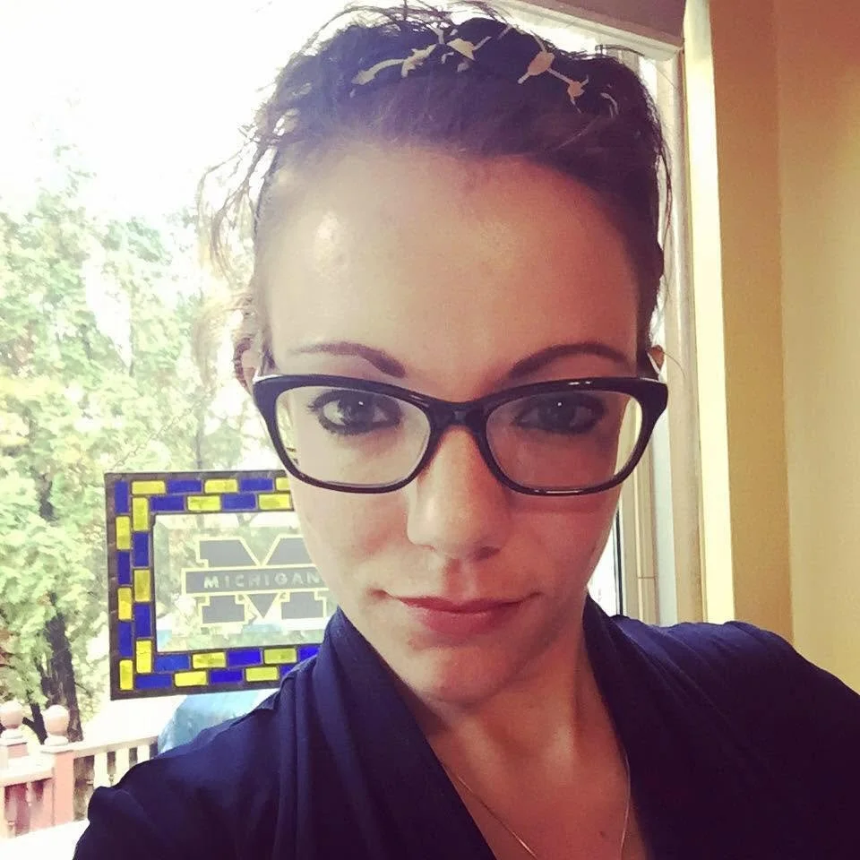 A woman with glasses and dark hair, taking a selfie indoors near a window with trees outside. There is a sign in the background with the word 'Michigan' and a block M logo.