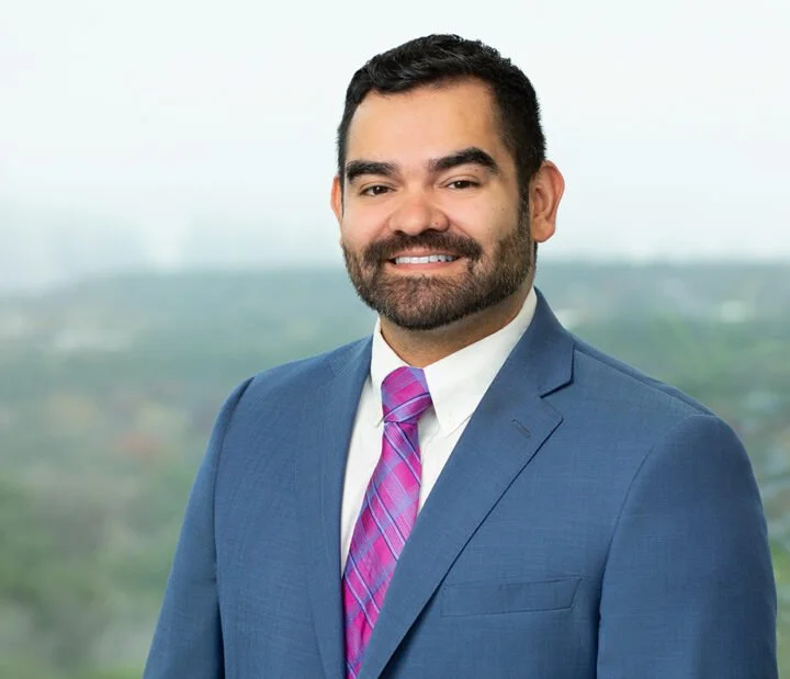 A man in a blue suit, white shirt, and purple pink striped tie standing outdoors with a blurred natural background.