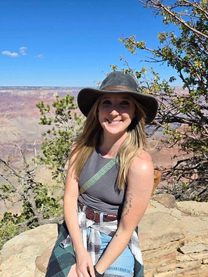 A smiling woman wearing a wide-brimmed hat, sleeveless gray top, and with a plaid shirt tied around her waist, standing on a rocky ledge at the Grand Canyon with trees and blue sky in the background.