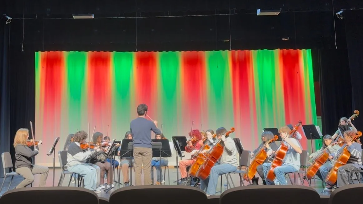 Young musicians in a string orchestra practicing under a colorful stage curtain with red, green, and yellow lighting, with a conductor leading.