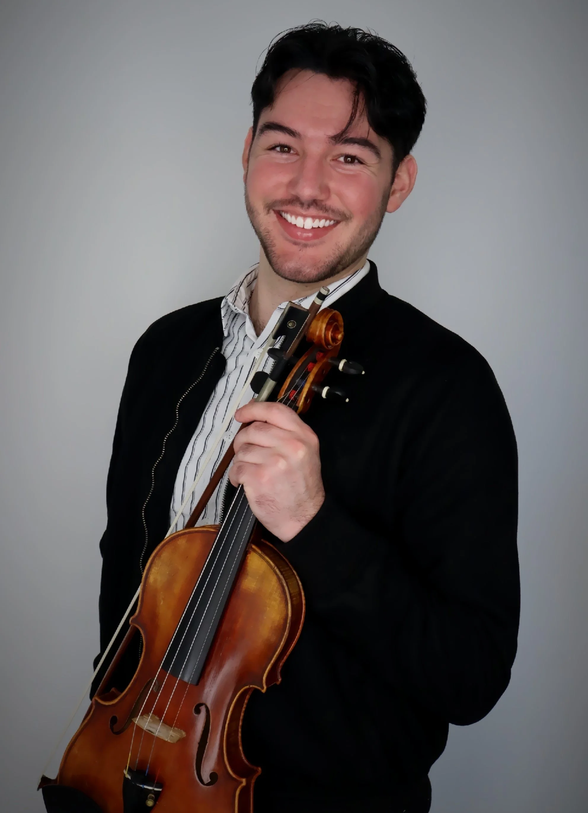 A man with black hair and a beard smiling while holding a violin in front of a plain gray background.