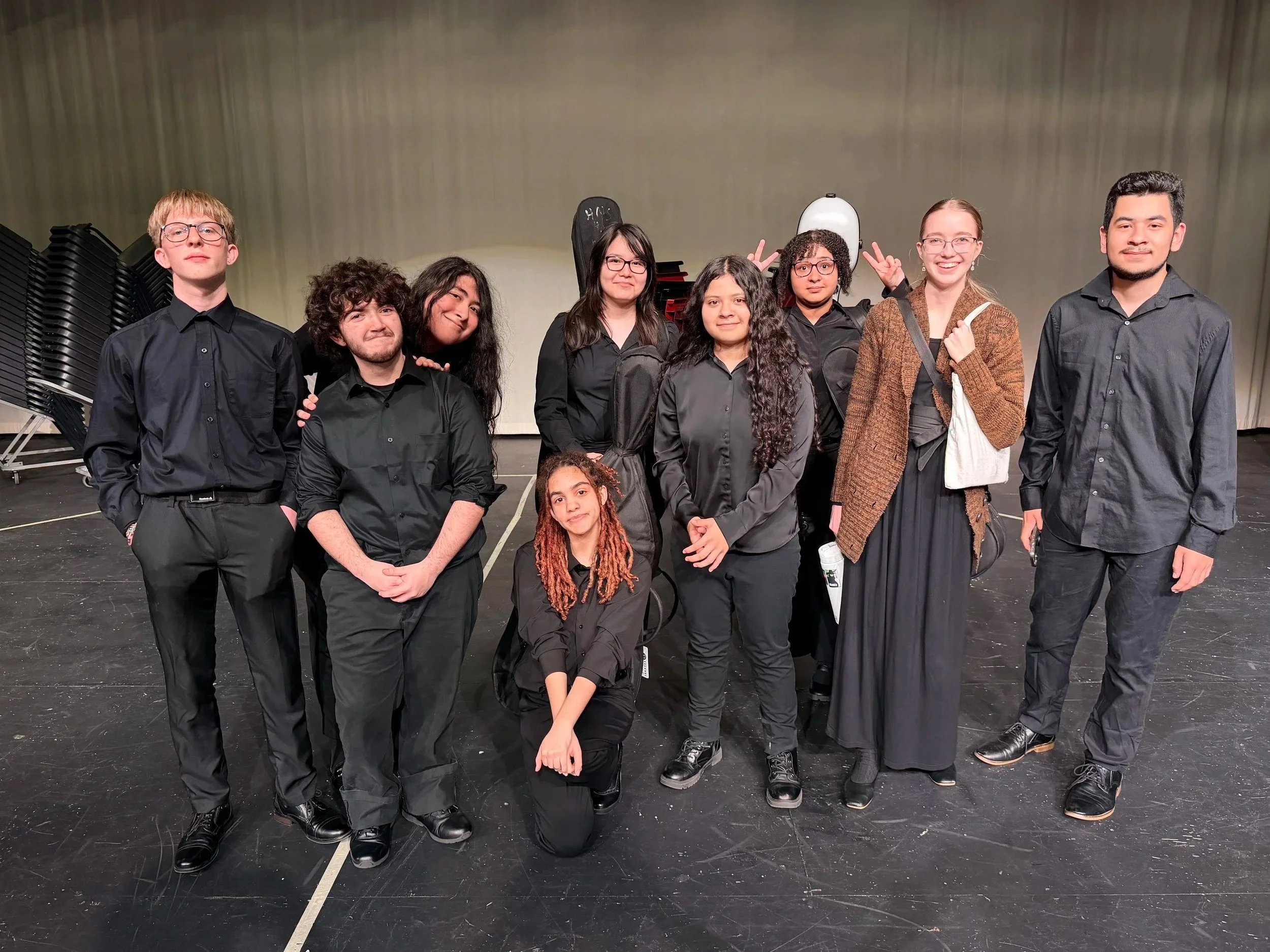 Group of nine young people, some making peace signs, dressed mostly in black, standing on a stage with black floor and a plain background, possibly after a performance.
