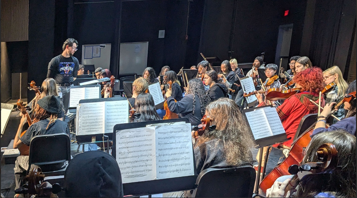 An orchestra rehearsing on stage, with musicians playing string instruments and a conductor leading them.