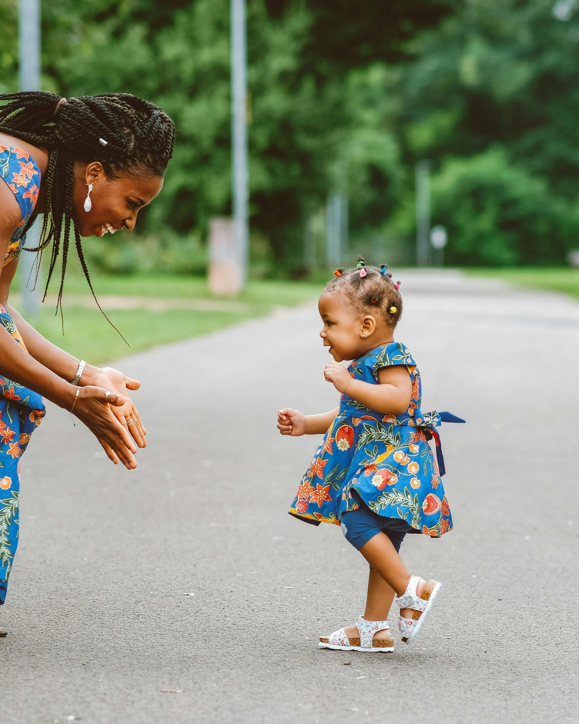 A woman and a young girl in matching floral dresses share a joyful moment outdoors. The girl is running toward the woman while smiling, and the woman is bending forward with her hands open, ready to catch her. They are on a paved path surrounded by greenery.