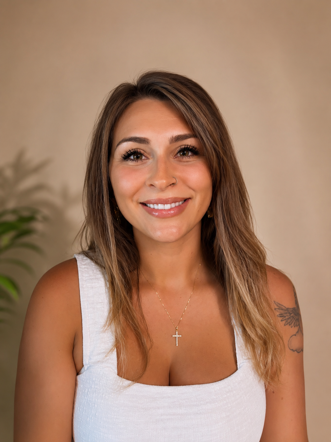 A smiling woman with long, wavy brown hair wearing a white sleeveless top, a gold cross necklace, and a small nose piercing, with a blurred green plant in the background.