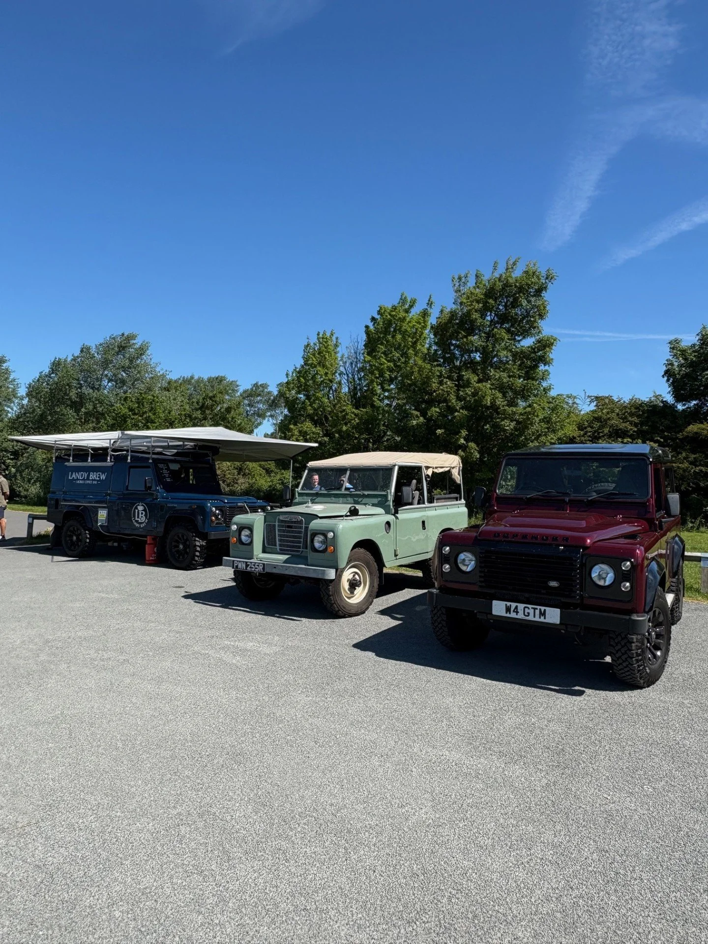 Three vintage Land Rover vehicles parked side by side on a gravel area under a blue sky with trees in the background. The vehicles are a dark blue Land Rover Defender, a light green Series Land Rover, and a maroon Land Rover Defender.