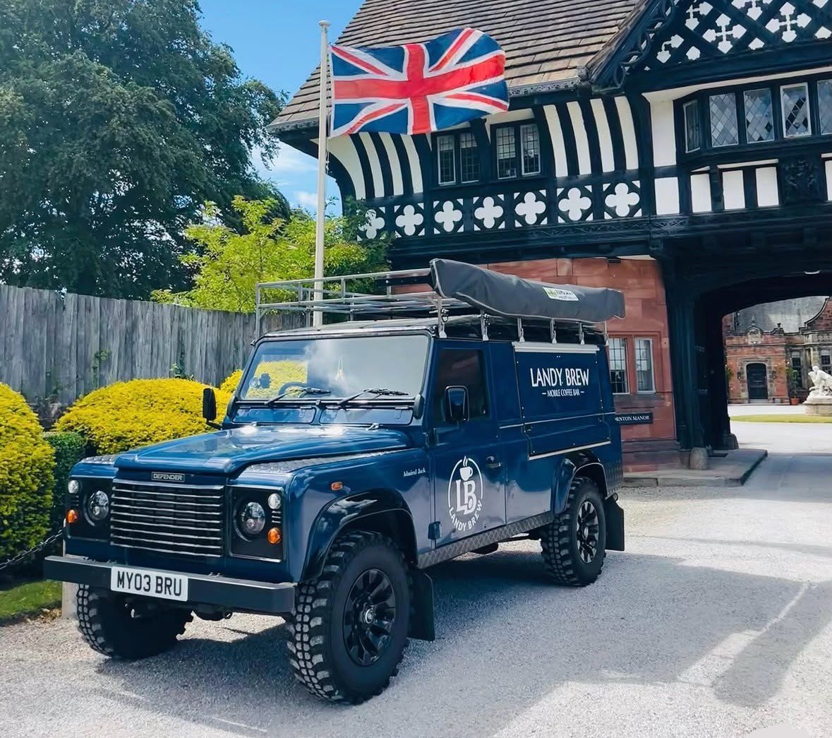 A blue Land Rover Defender vehicle with branding for Landy Brew Mobile Coffee, parked in front of a traditional black-and-white Tudor-style building, with a Union Jack flag on a pole flying above.