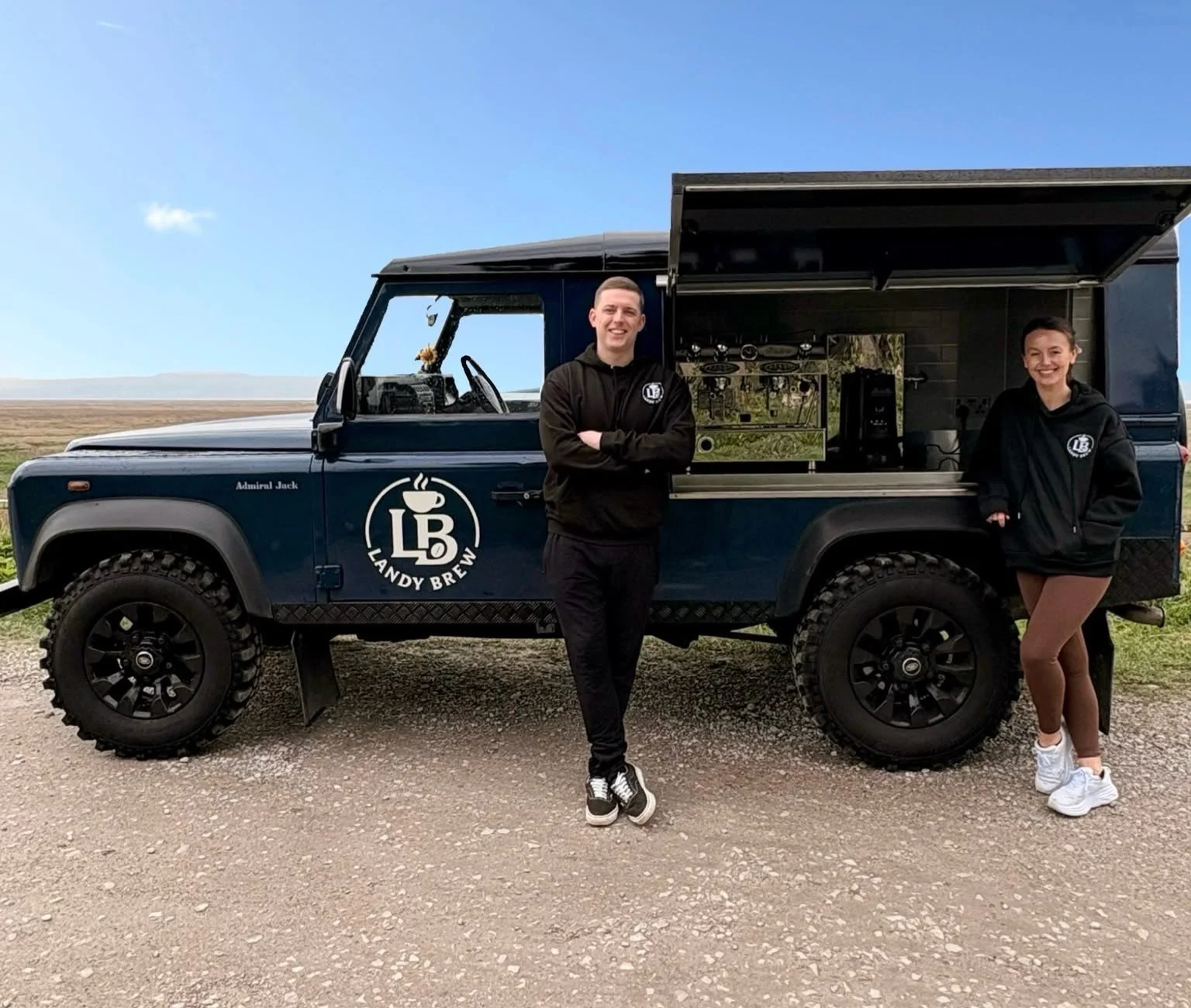 Two people standing in front of a blue Landy Brew mobile coffee truck. The man on the left is wearing a black hoodie and black pants with sneakers, smiling with arms crossed. The woman on the right is wearing a black hoodie, brown pants, and white sneakers, smiling with hands in hoodie pockets. The coffee truck has a logo with a coffee cup and the words "LB Landy Brew" on the side, with a window open showing coffee equipment inside. The background features a flat, open landscape with a blue sky.