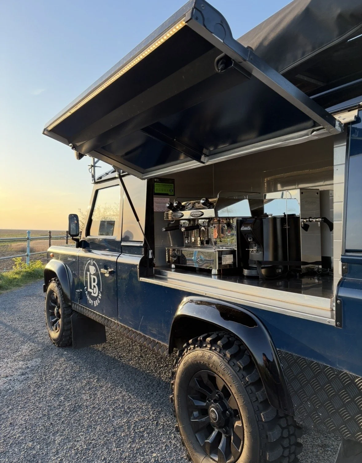 A blue food truck with a service window open, featuring coffee machines and equipment inside, parked outdoors during sunset.