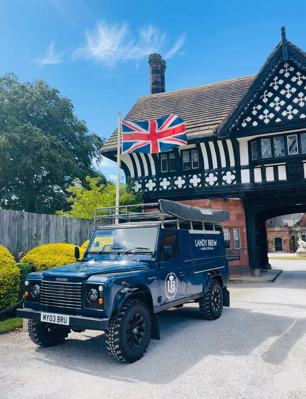 A dark blue Land Rover Defender parked in front of a Tudor-style building with black and white timber framing, a red brick chimney, and a wooden fence. A Union Jack flag is flying on a flagpole next to the vehicle, and there are green bushes and tree