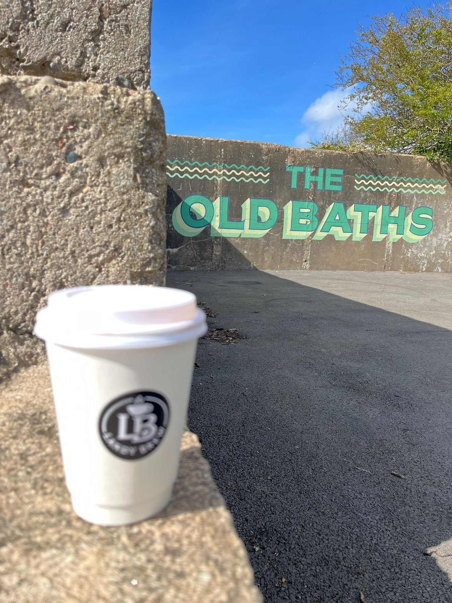 A paper coffee cup with a logo in the foreground, a stone wall on the left, and a concrete wall with painted text saying "The Old Baths" in the background, under a blue sky with some trees.