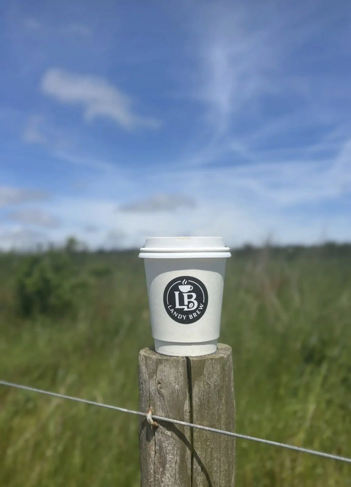 A white coffee cup with lid, labeled 'Landy Brew' and a logo, placed on a wooden fence post in a grassy outdoor area with a blue sky and clouds in the background.