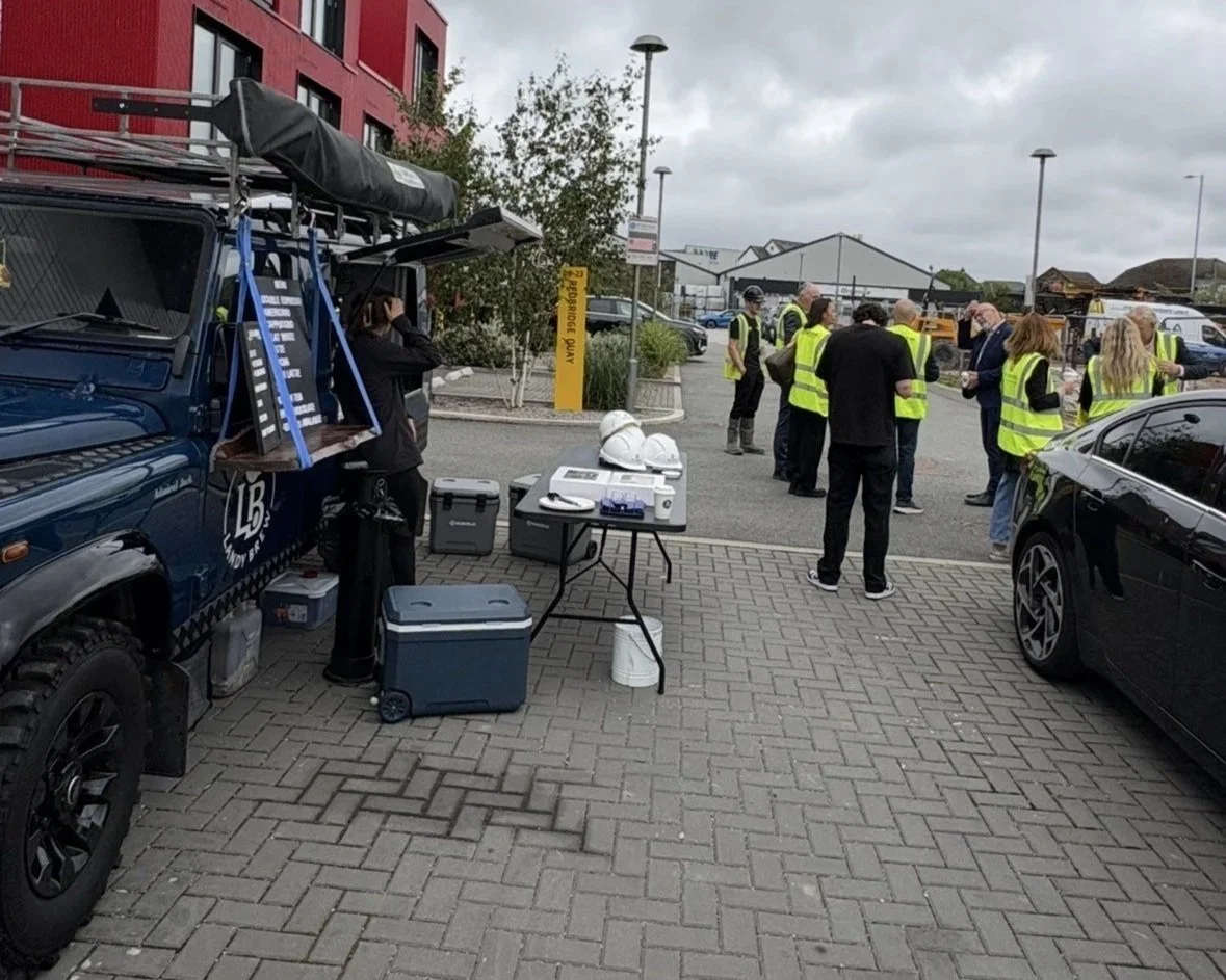 Group of people in yellow safety vests gathered outdoors near parked cars, with a table holding safety helmets and equipment, and a work vehicle nearby under cloudy skies.