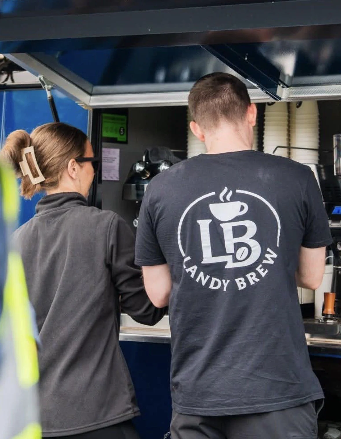 Two people working behind a coffee stand. One person is wearing a black T-shirt with a coffee cup and 'LAB' logo, and 'Candy Brew' text on the back. The other person is wearing a dark jacket and glasses.