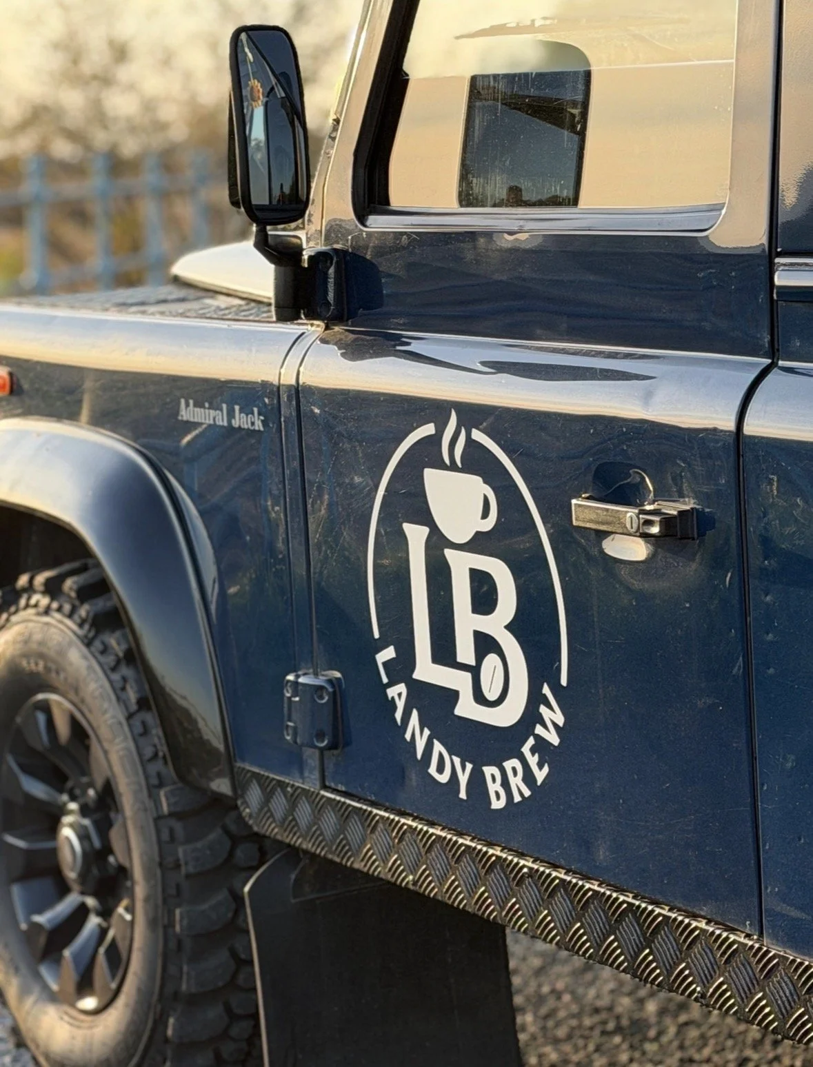 Close-up of a blue off-road vehicle with a Landy Brew logo that includes a coffee cup and the letters 'LB'. The vehicle has a side mirror, rugged tires, and a decorative metal step.