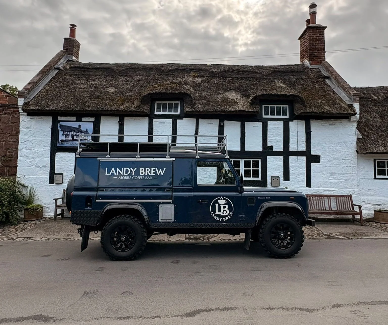 A vintage black Land Rover with a sign that reads 'Landy Brew Mobile Coffee Bar' parked in front of a traditional white cottage with a thatched roof and black timber framing, on a cobblestone pathway.