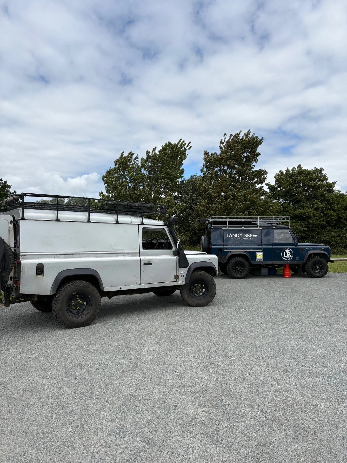 Two parked vehicles, one white and one dark blue, are in an open area with a cloudy sky and trees in the background. The blue vehicle has a sign for a mobile coffee bar called 'Landy Brew'.