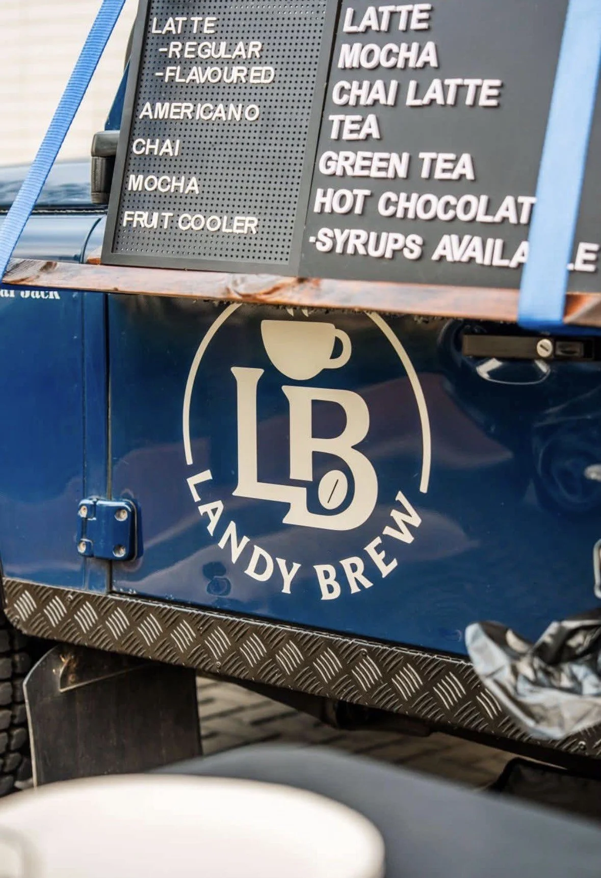 A blue food truck with a menu sign listing various coffee drinks and a logo reading Les Landing Brew.