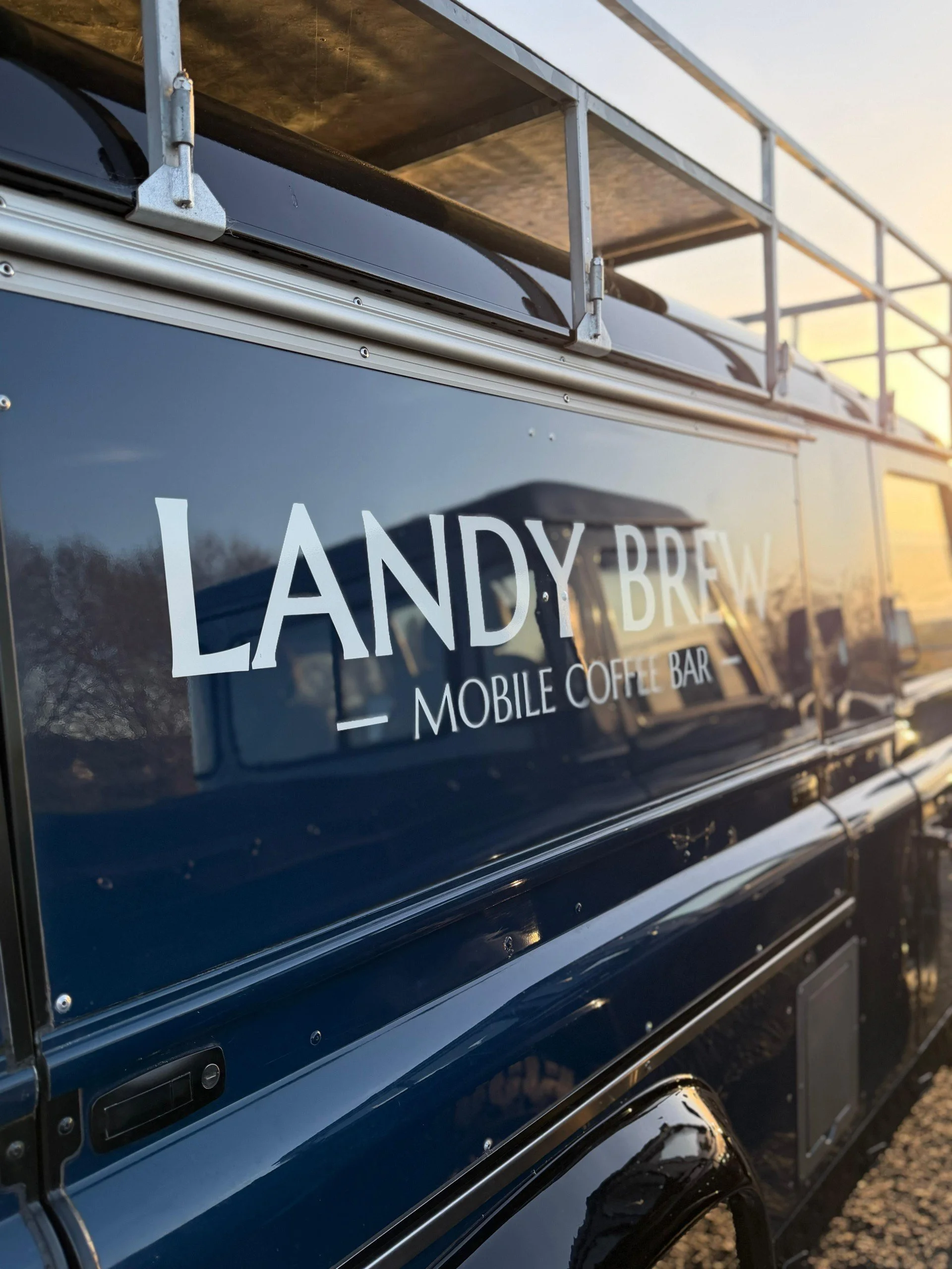A blue vehicle with a sign reading 'LANDY BREW' on its side, indicating it is a mobile coffee bar. The vehicle has a metal railing or canopy on top and is parked outdoors during sunset.